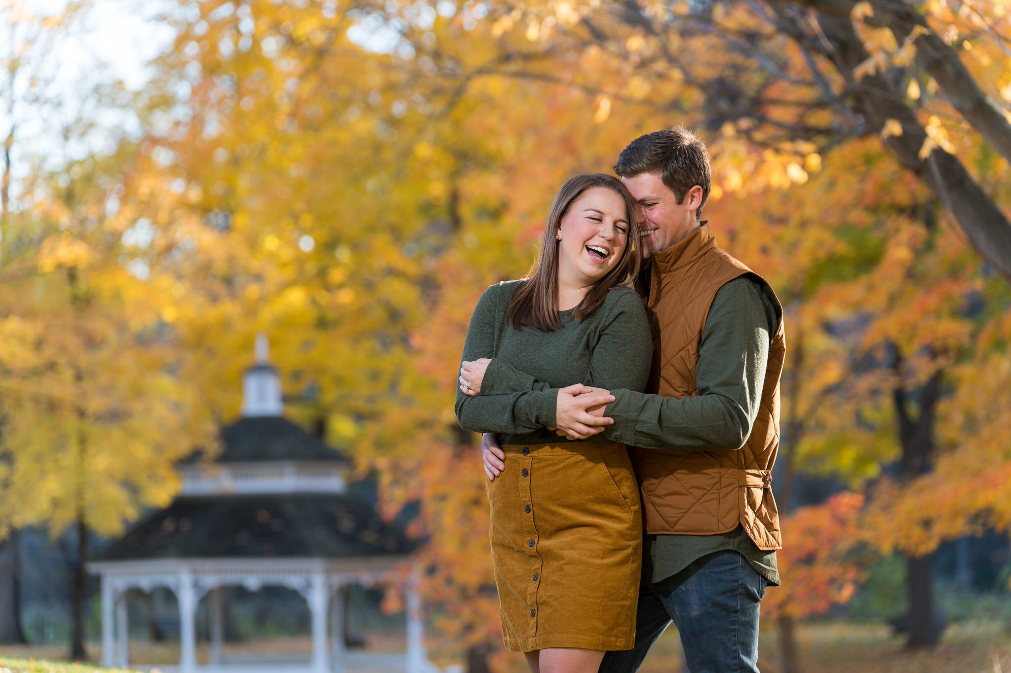 A man hugs his fiance from behind and laughs at a fall engagement at Van Hoosen. 