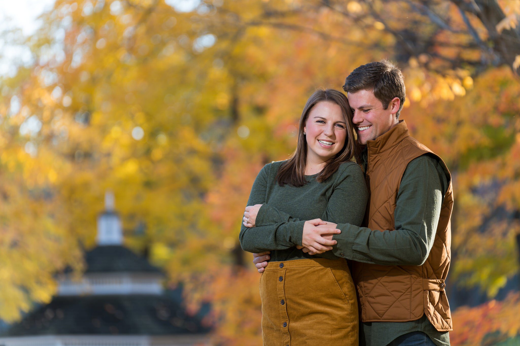 A man hugs his fiance from behind and smiles at a fall engagement at Van Hoosen. 