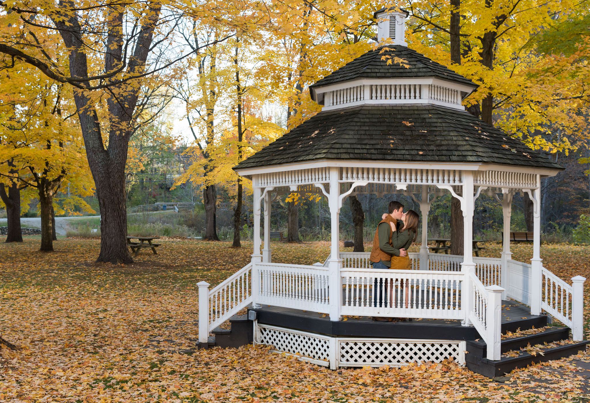 Standing in a white gazebo, a couple kisses at their fall engagement at Van Hoosen. 