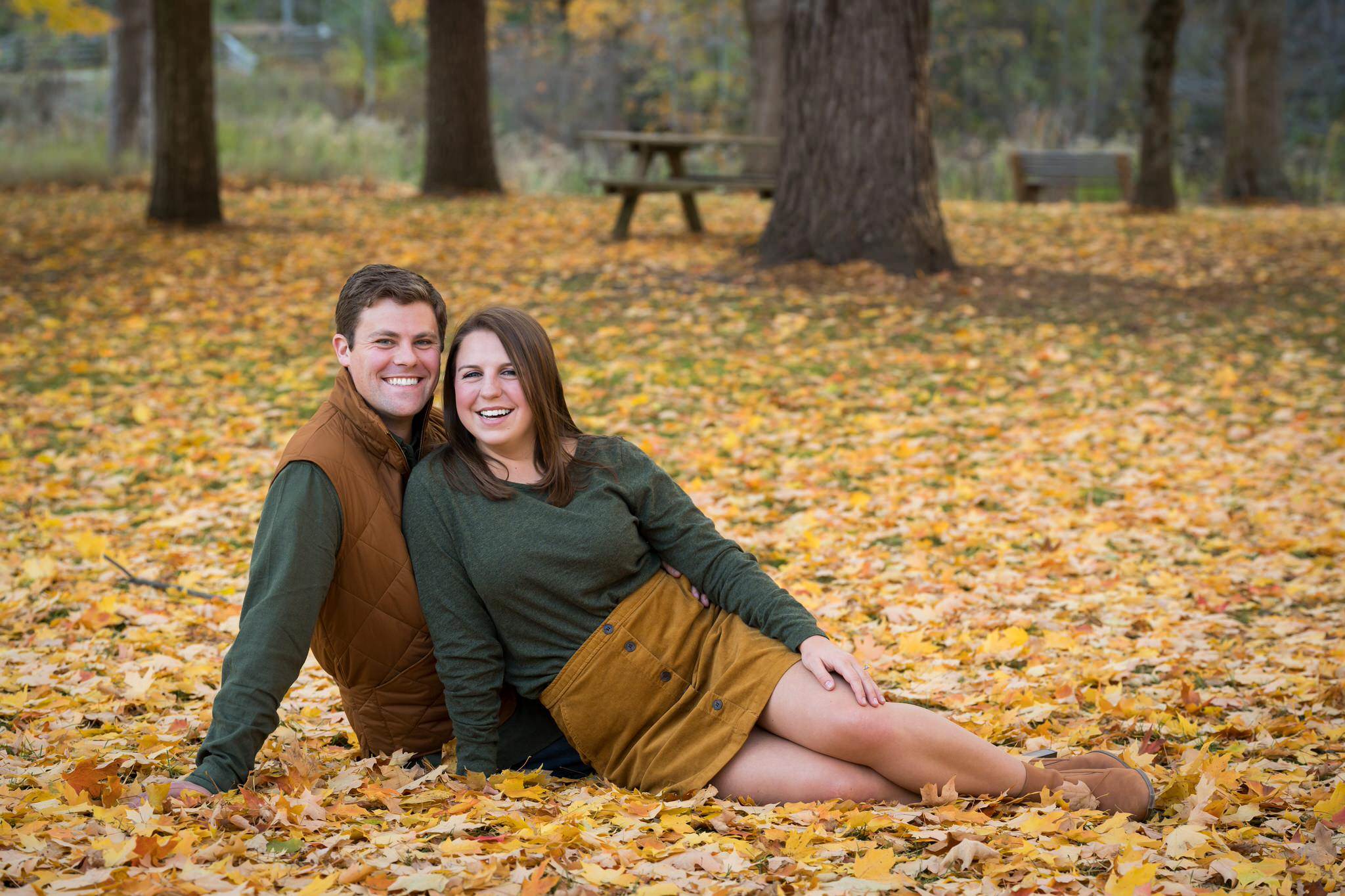 A couple, wearing green shirts, sits on the ground and smiles during their  fall engagement at Van Hoosen. 