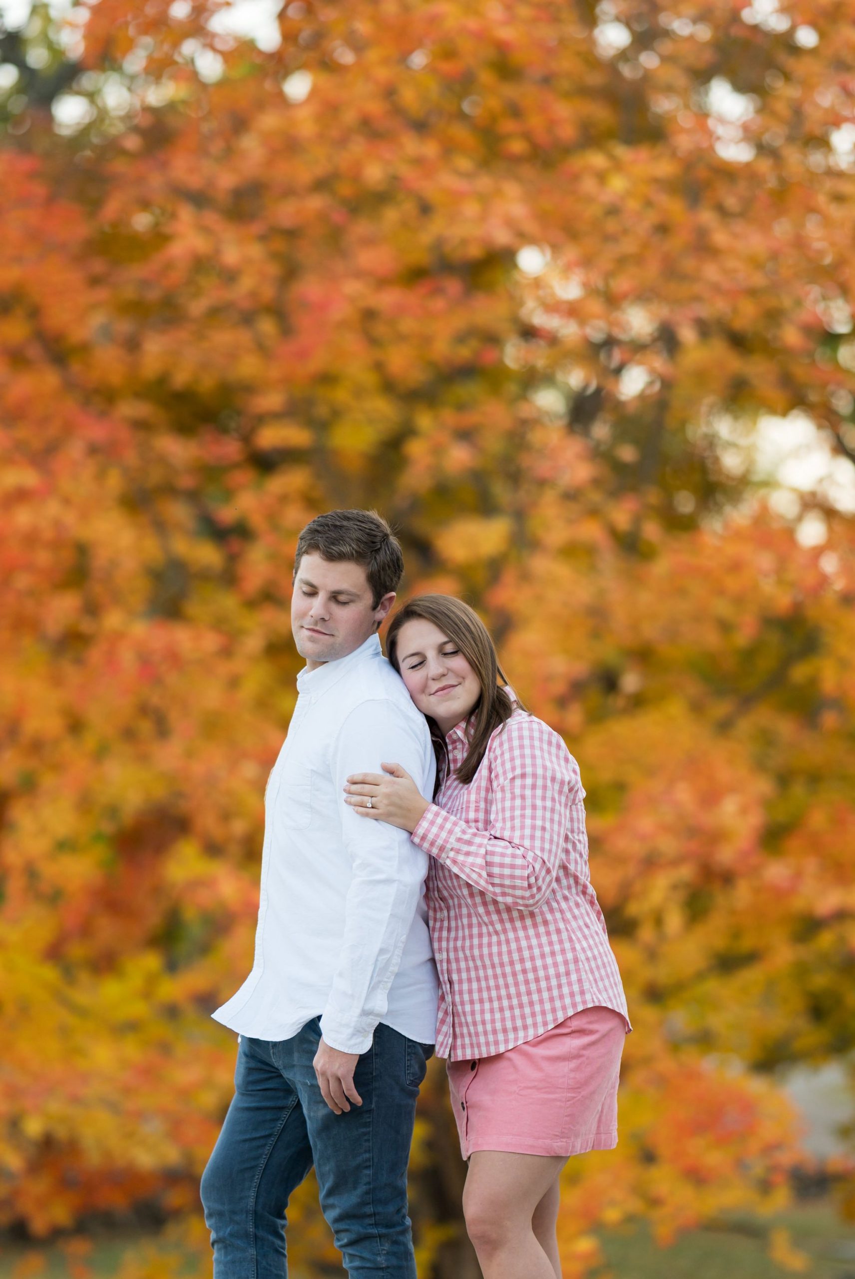 A woman hugs her fiance from behind with the backdrop of an orange leafed tree behind them at their fall engagement at Van Hoosen. 