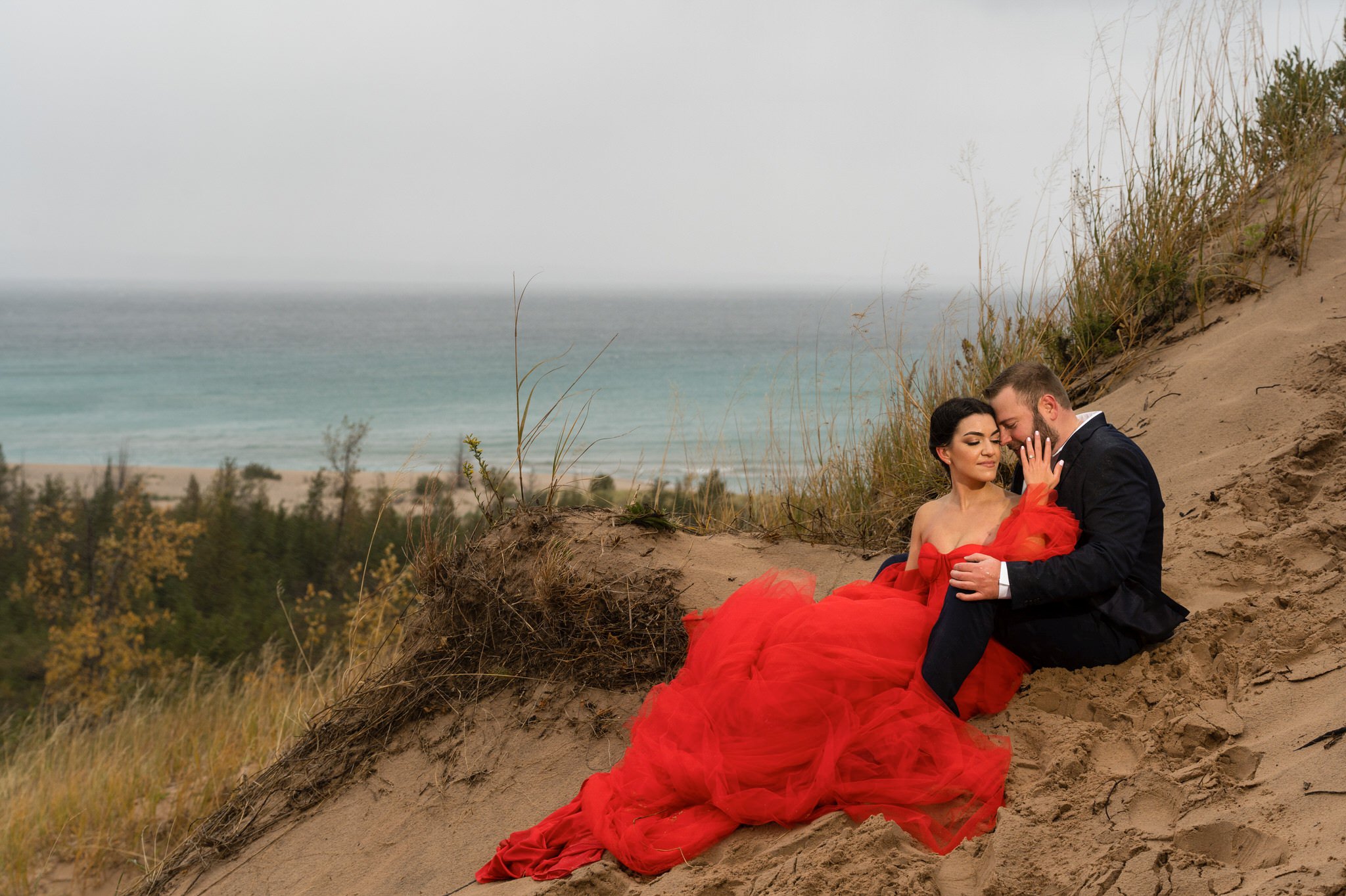 A couple sits and cuddles on a sand dune with grey clouds in the background at Sleeping Bear Dunes engagement session.