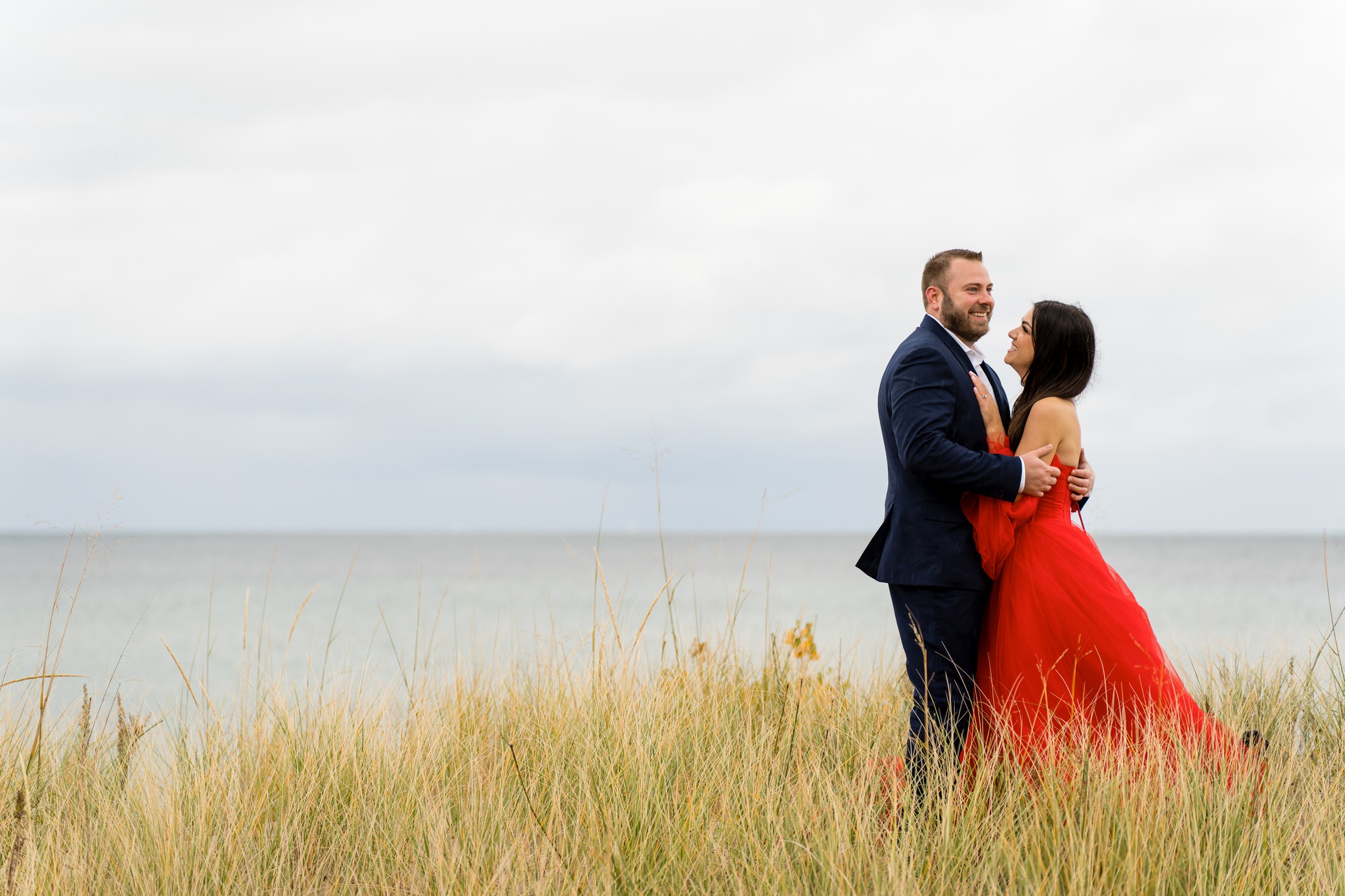 Standing in tall grass near the shoreline, a couple hugs during their Sleeping Bear Dunes engagement session. 