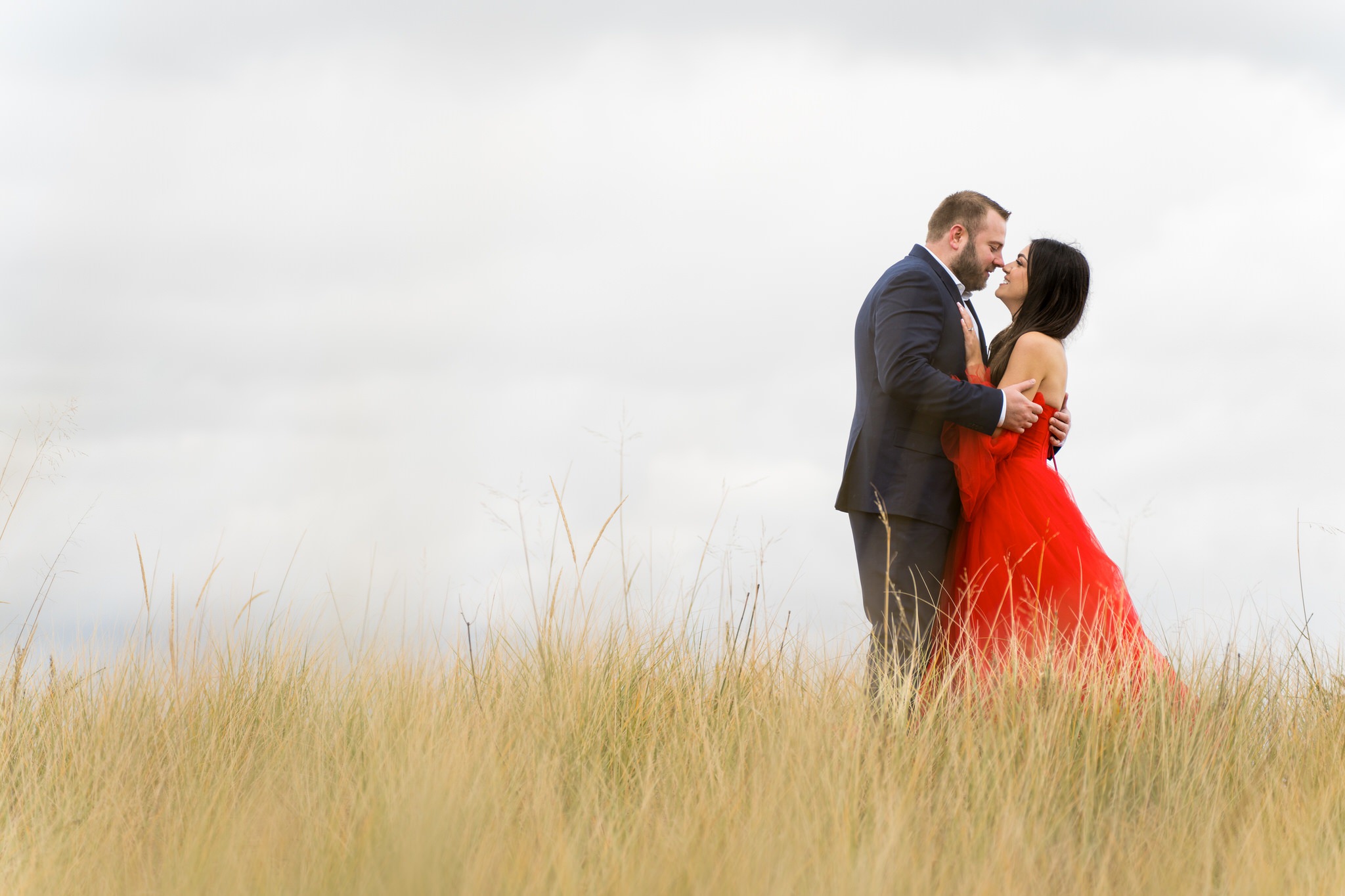 Standing in tall grass near the shoreline, a couple hugs during their Sleeping Bear Dunes engagement session. 