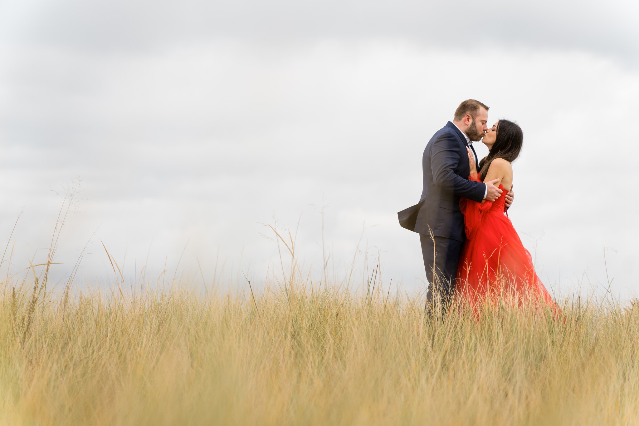 Standing in tall grass near the shoreline, a couple kisses during their Sleeping Bear Dunes engagement session. 