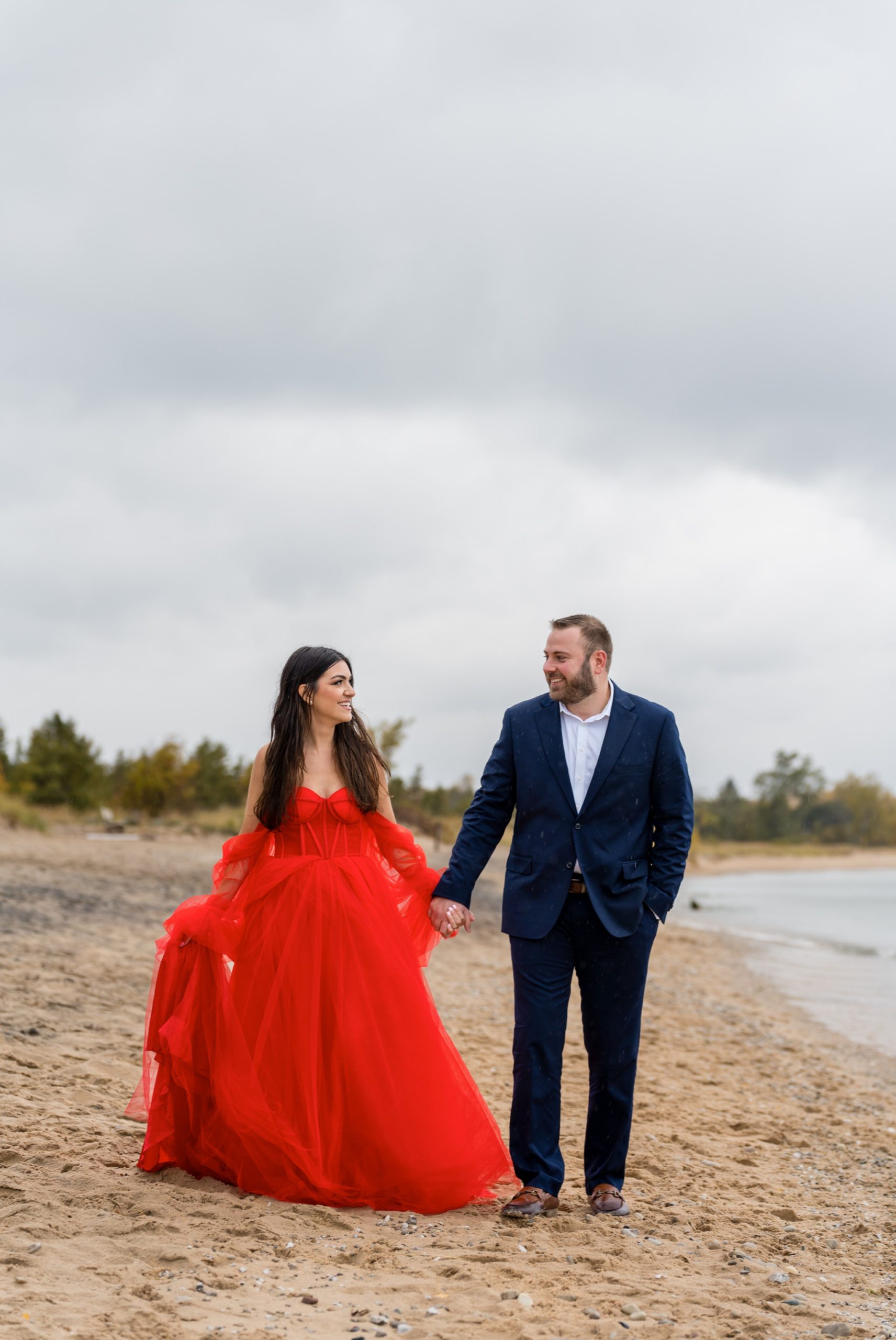 A couple, wearing a flowing red dress and blue suit, walk the shoreline during their Sleeping Bear Dunes engagement session.