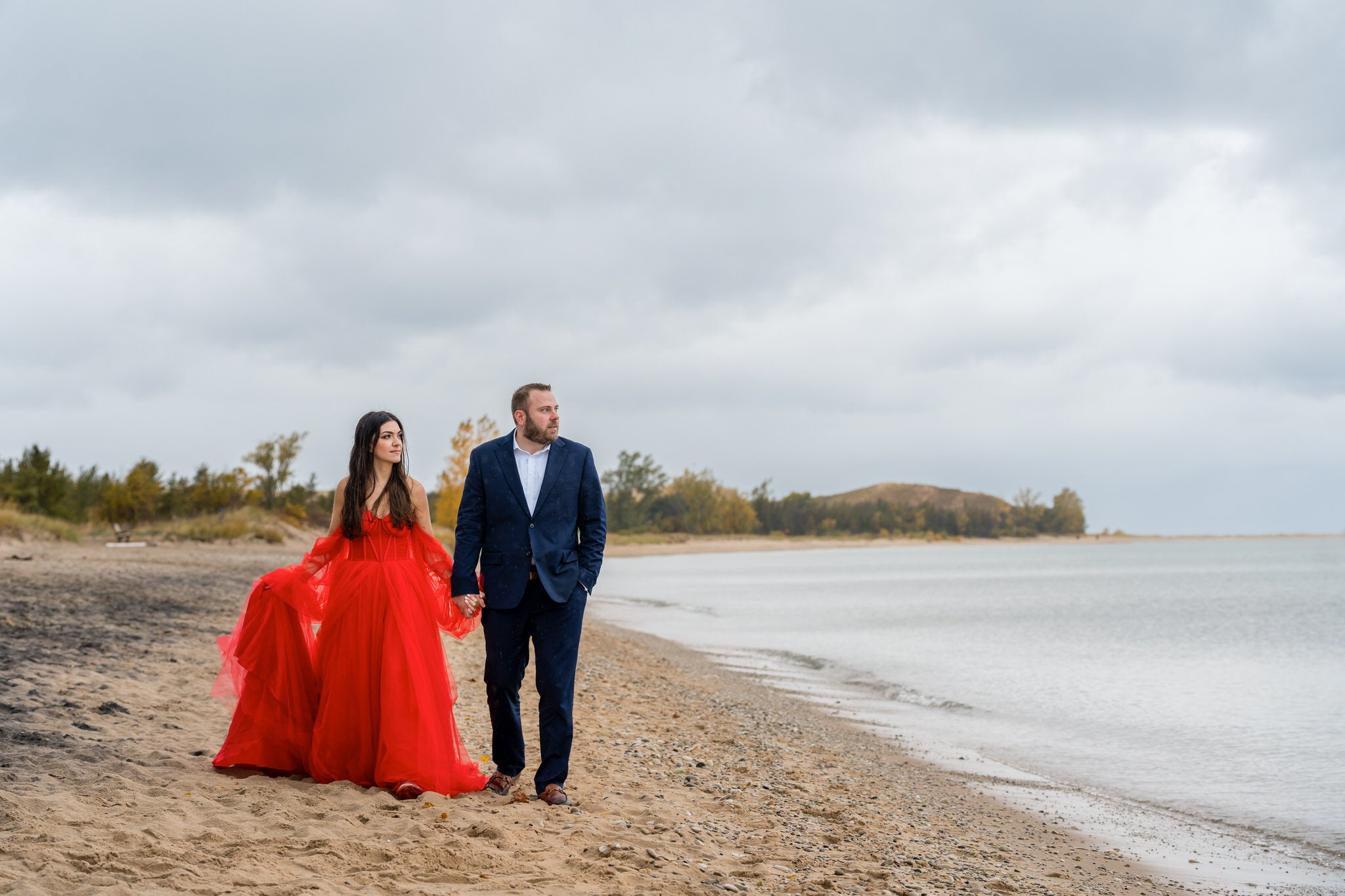 A couple, wearing a flowing red dress and blue suit, walk the shoreline during their Sleeping Bear Dunes engagement session.