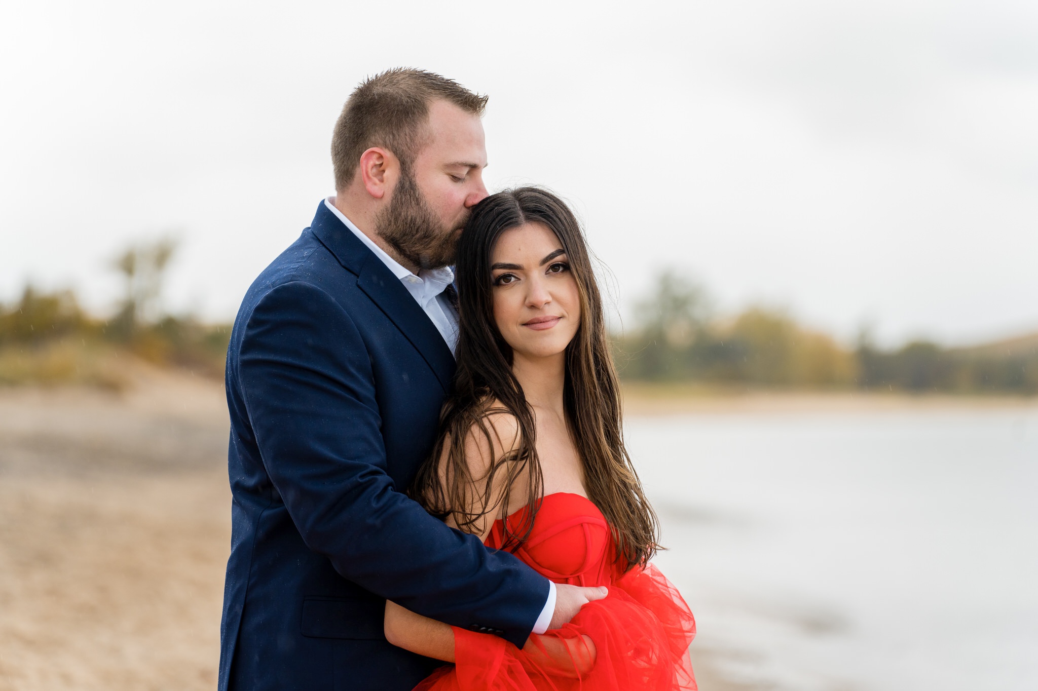 A couple, wearing a flowing red dress and blue suit, hug at the shoreline during their Sleeping Bear Dunes engagement session.