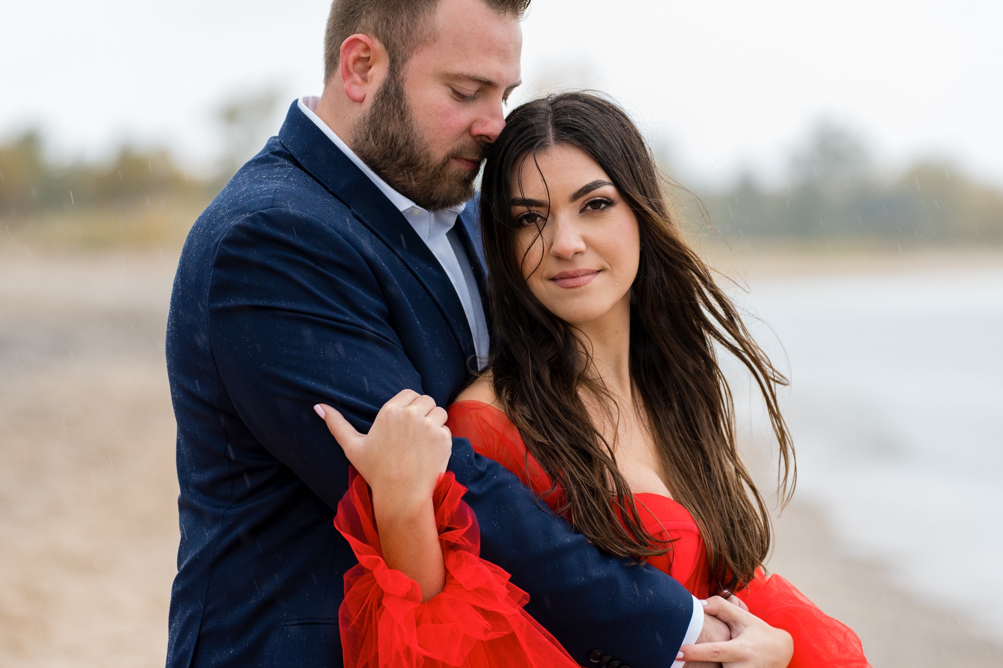 A couple, wearing a flowing red dress and blue suit, hug at the shoreline during their Sleeping Bear Dunes engagement session.