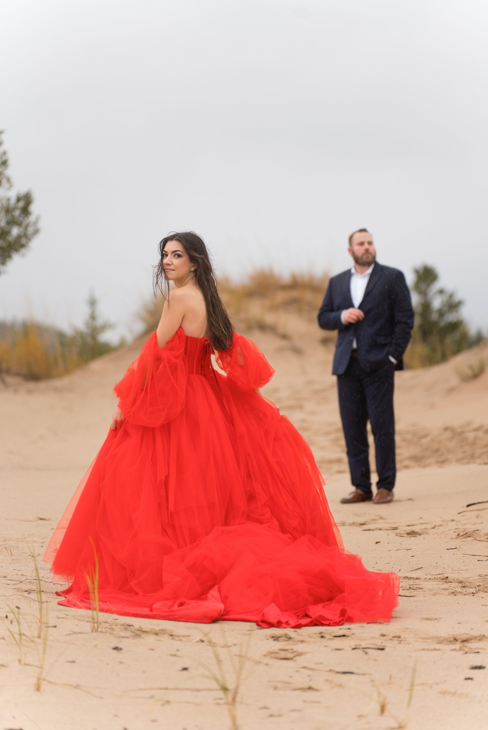 A full length photo of a woman, wearing a red flowing dress, looks over her shoulder at Sleeping Bear Dunes engagement session. 