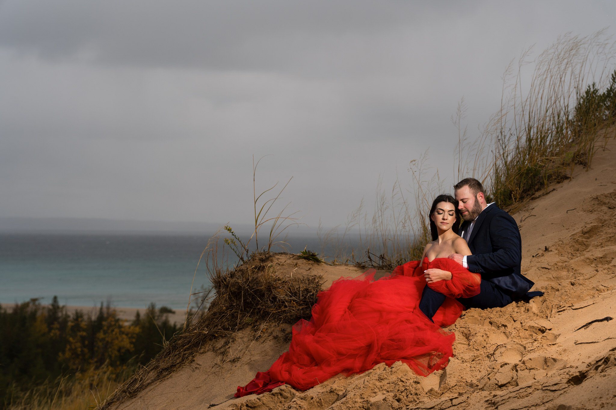 A couple sits and cuddles on a sand dune with grey clouds in the background at Sleeping Bear Dunes engagement session.
