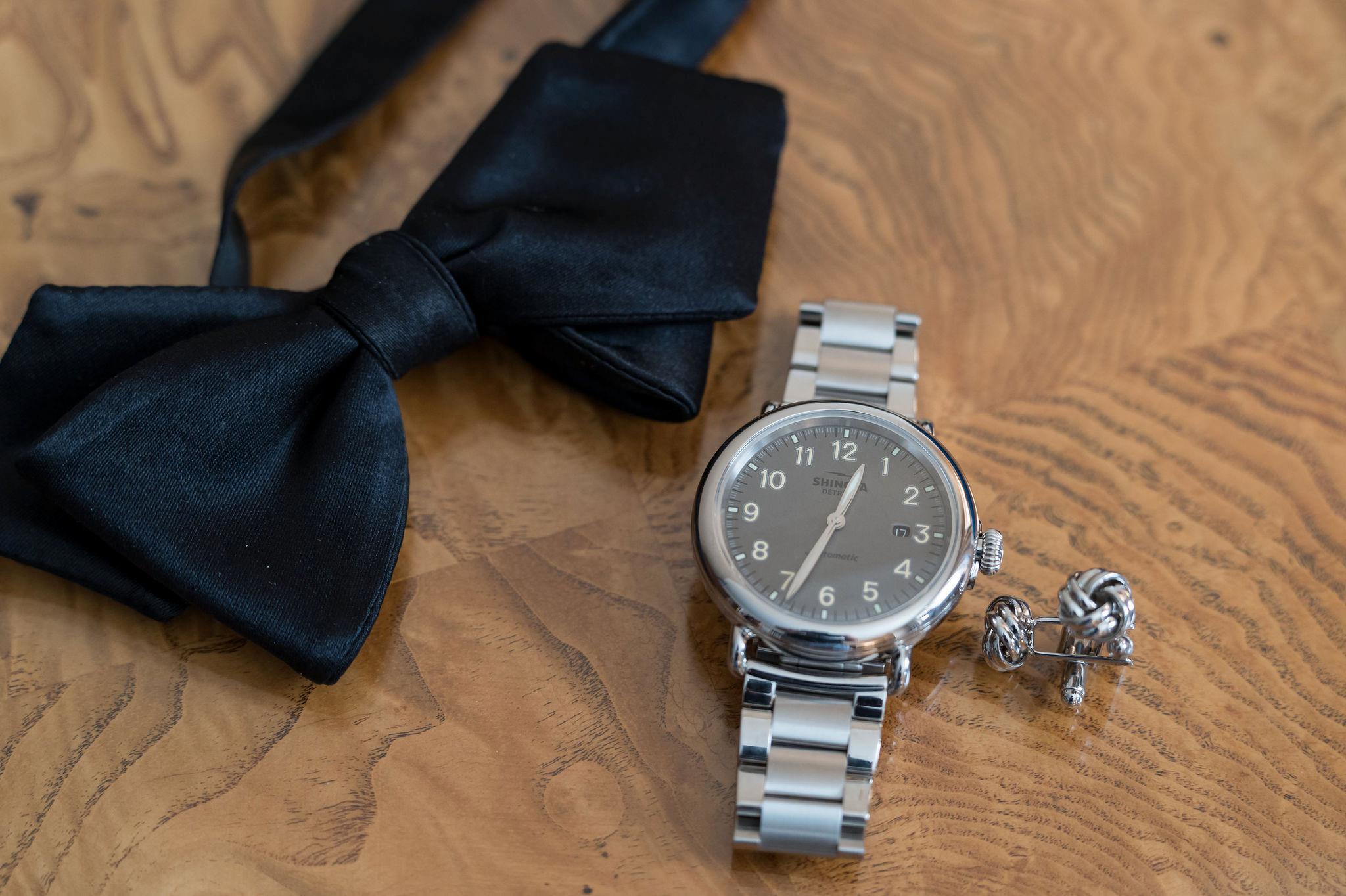A Shinola watch sits on a wood desk with cufflinks and black bowtie.