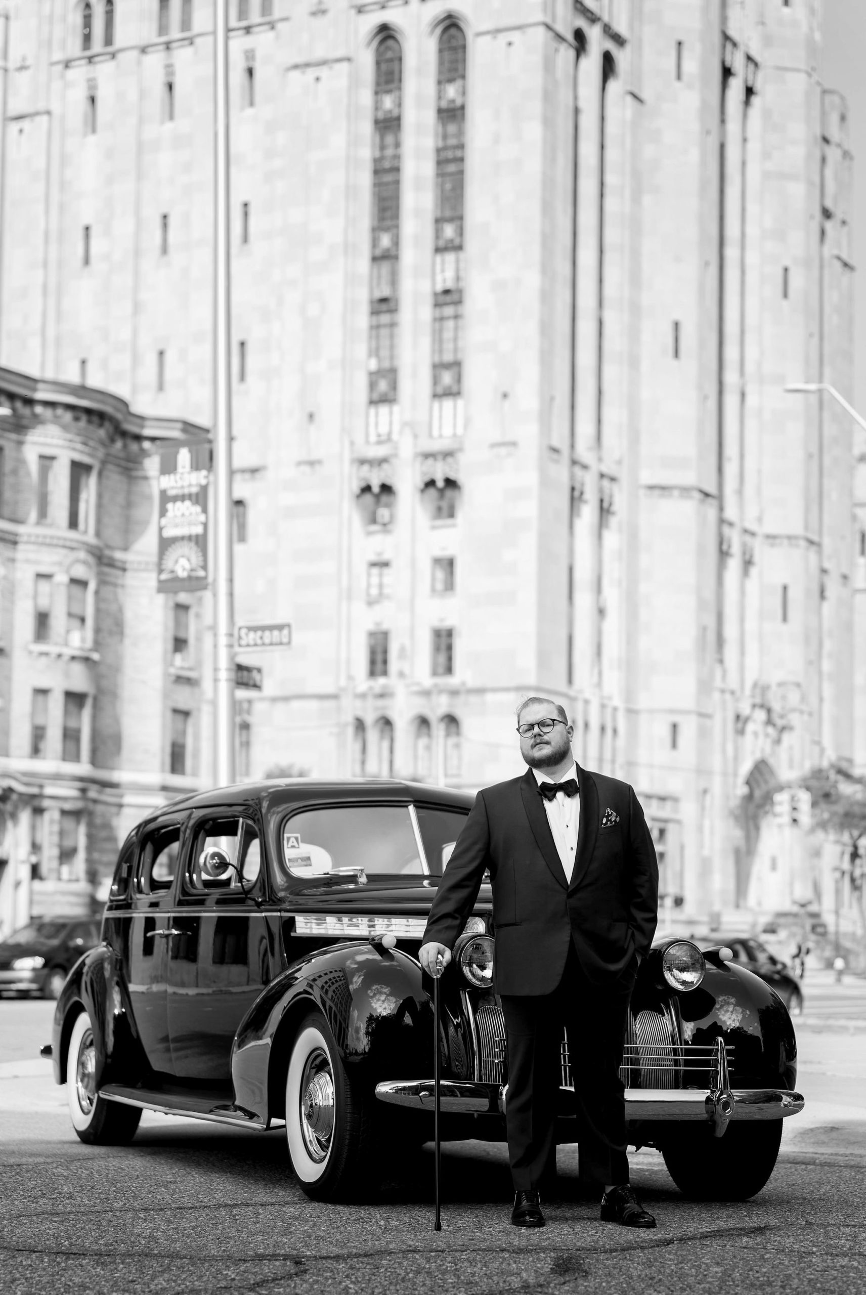 A groom poses with a classic car on the streets of Detroit near the Masonic Temple.