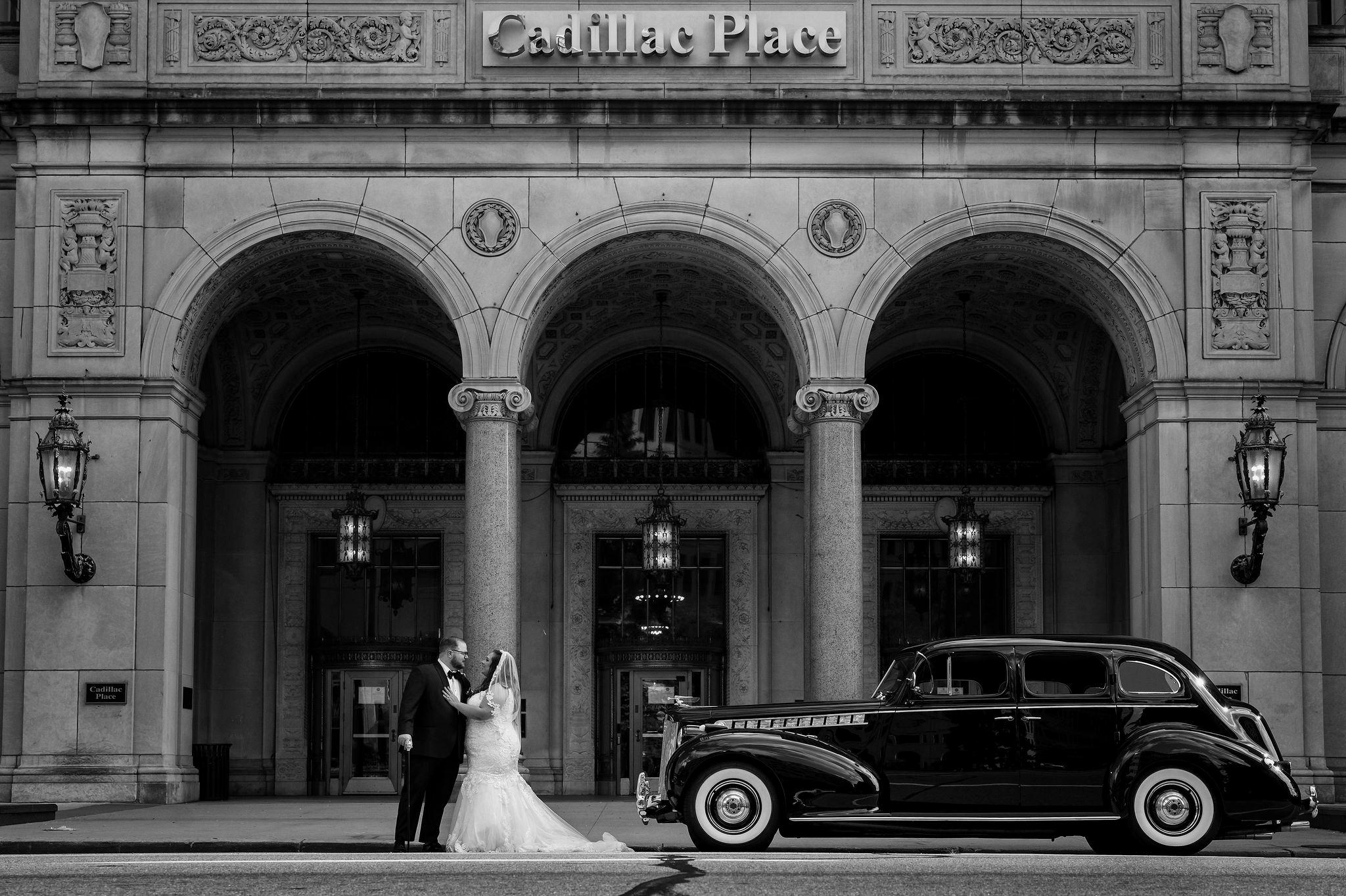 A couple poses with a classic car on the streets of Detroit in front of Cadillac Place Apartments.
