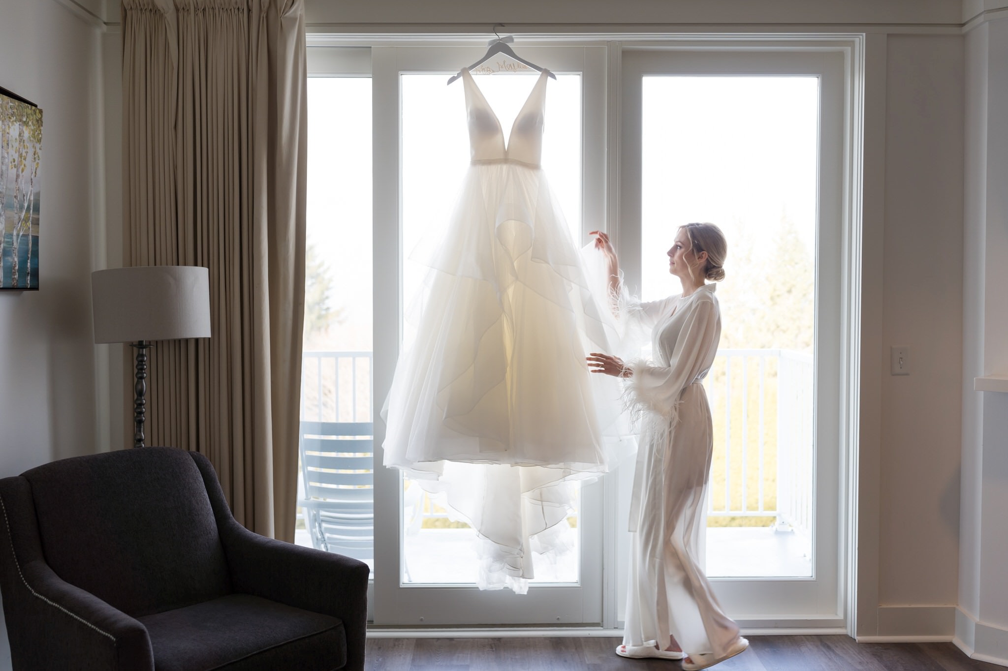 A bride, dressed in a white robe, approaches her wedding dress as it hangs by a window on her Bay Harbor wedding day.  