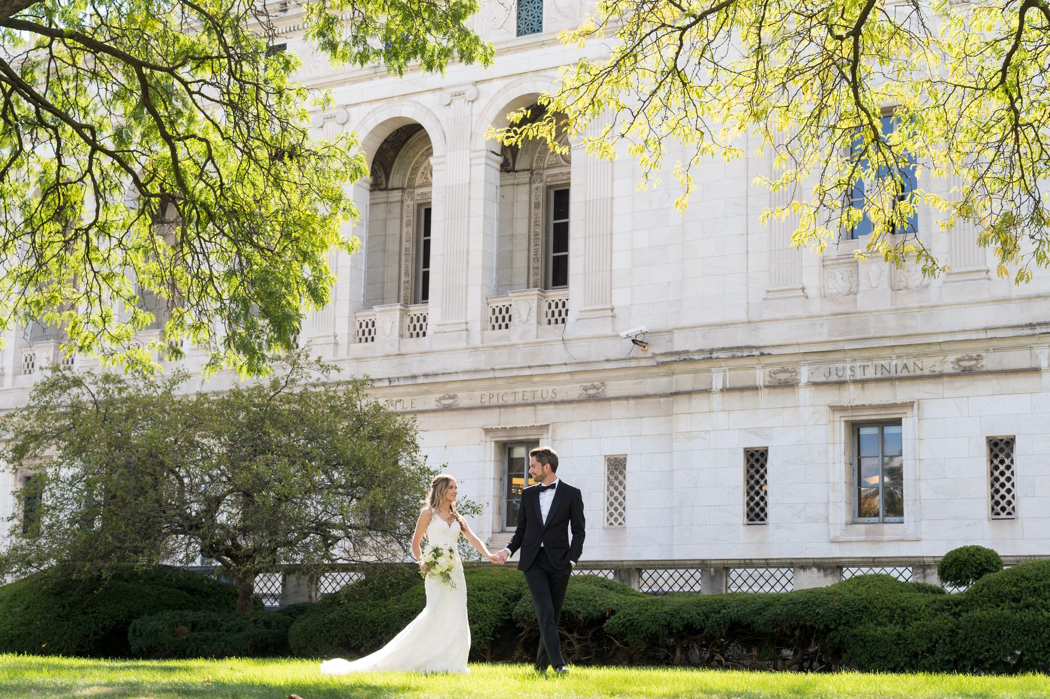 A bride and groom hold hands and walk across the grounds of the Detroit Public Library.