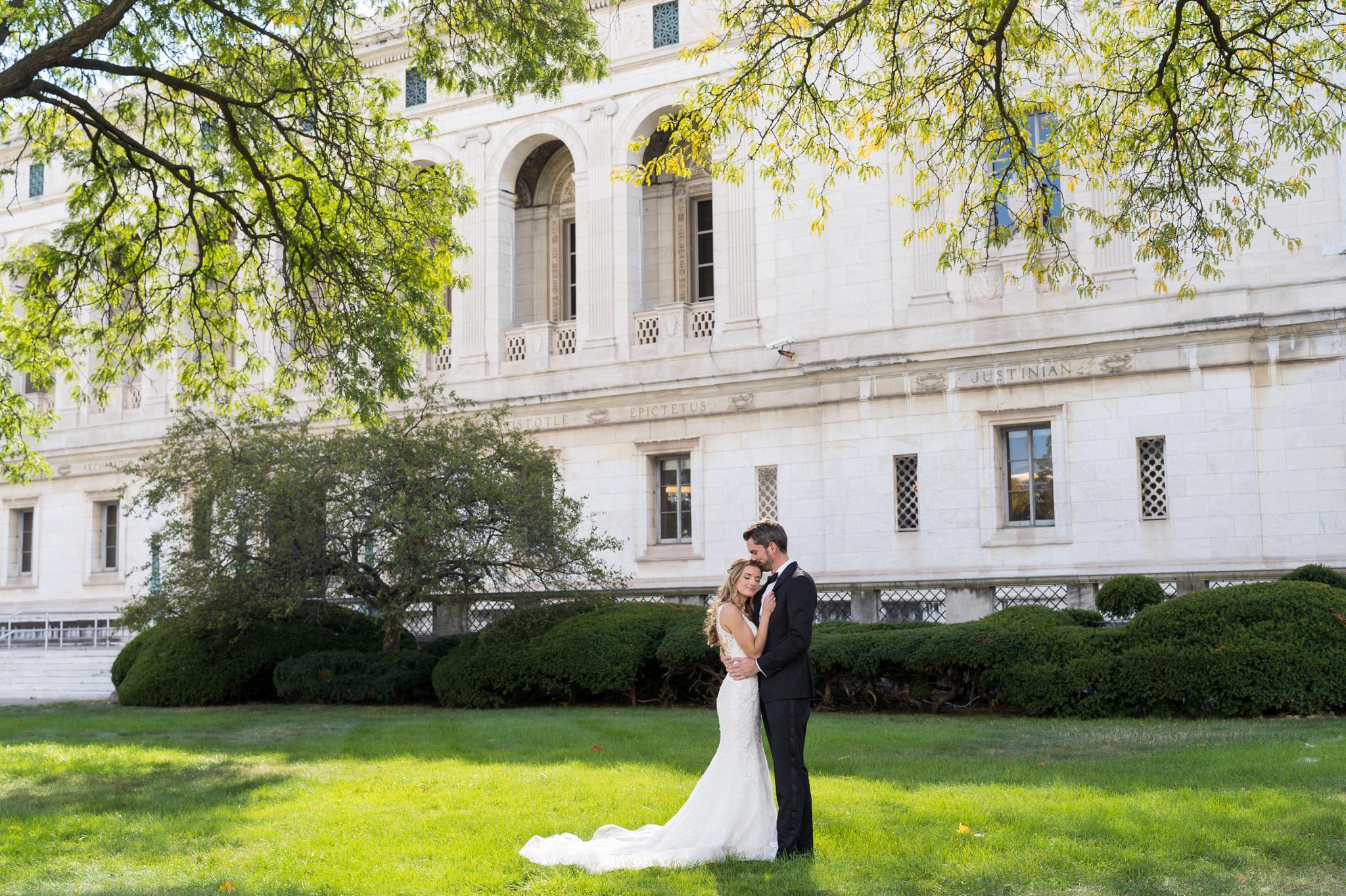 A groom gently kisses his brides head  standing outside of the Detroit Public Library.