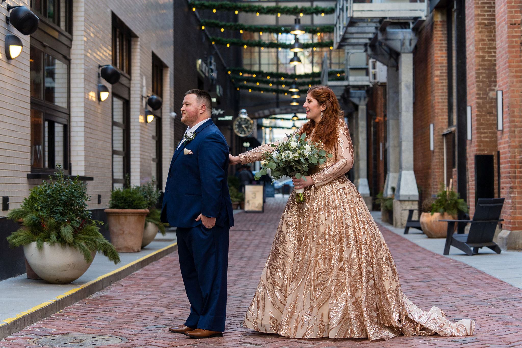 A bride, wearing a gold wedding dress, taps her groom on the shoulder during their first look in Parker's Alley.  
