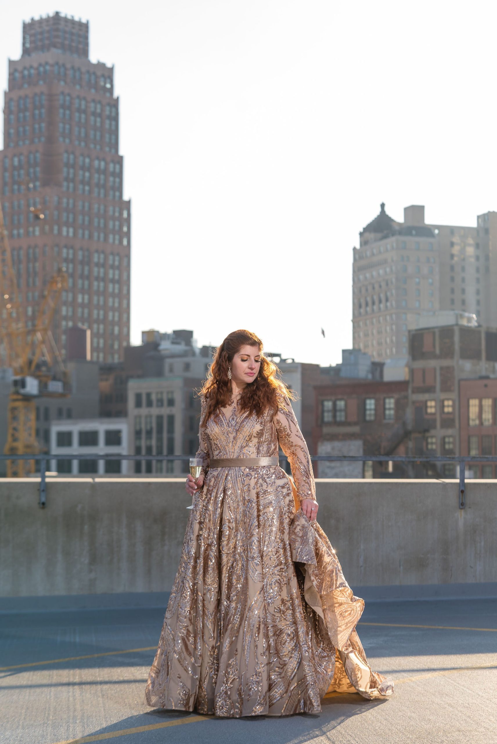 A bride, wearing a gold dress and holding a champagne glass, poses with Detroit skyline in the background. 