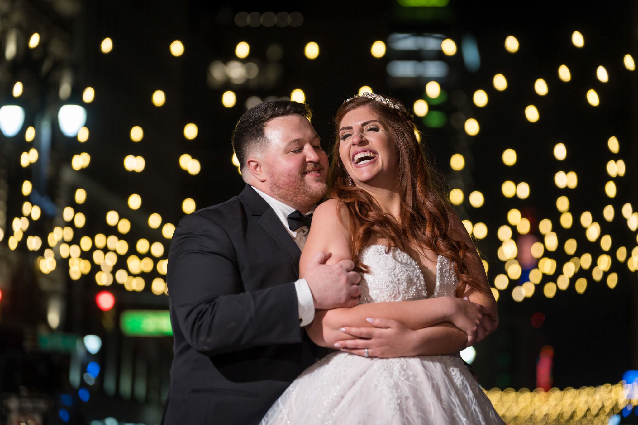 A groom hugs his bride from behind with twinkle lights overhead on Woodward Avenue in Detroit, Michigan.