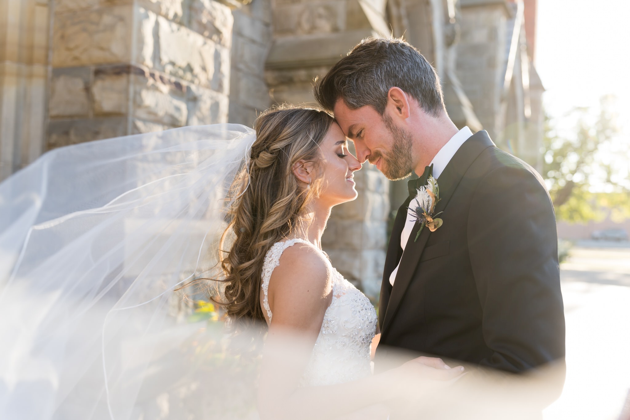 A bride and groom stand forehead to forehead as her veil blows in the wind, passing in front of the camera.