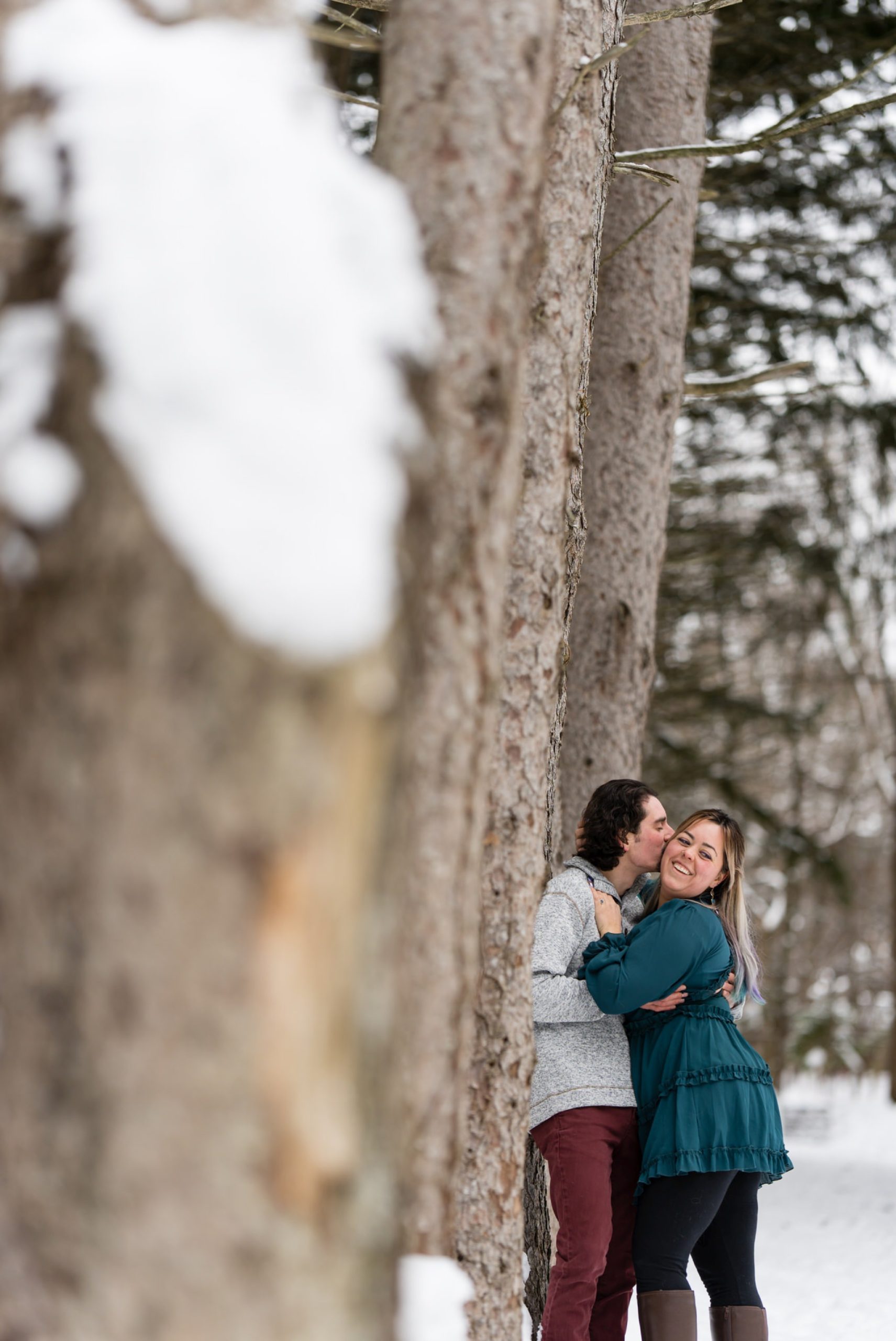 A couple pose near a tree in the snow for their winter engagement session at Stony Creek. 