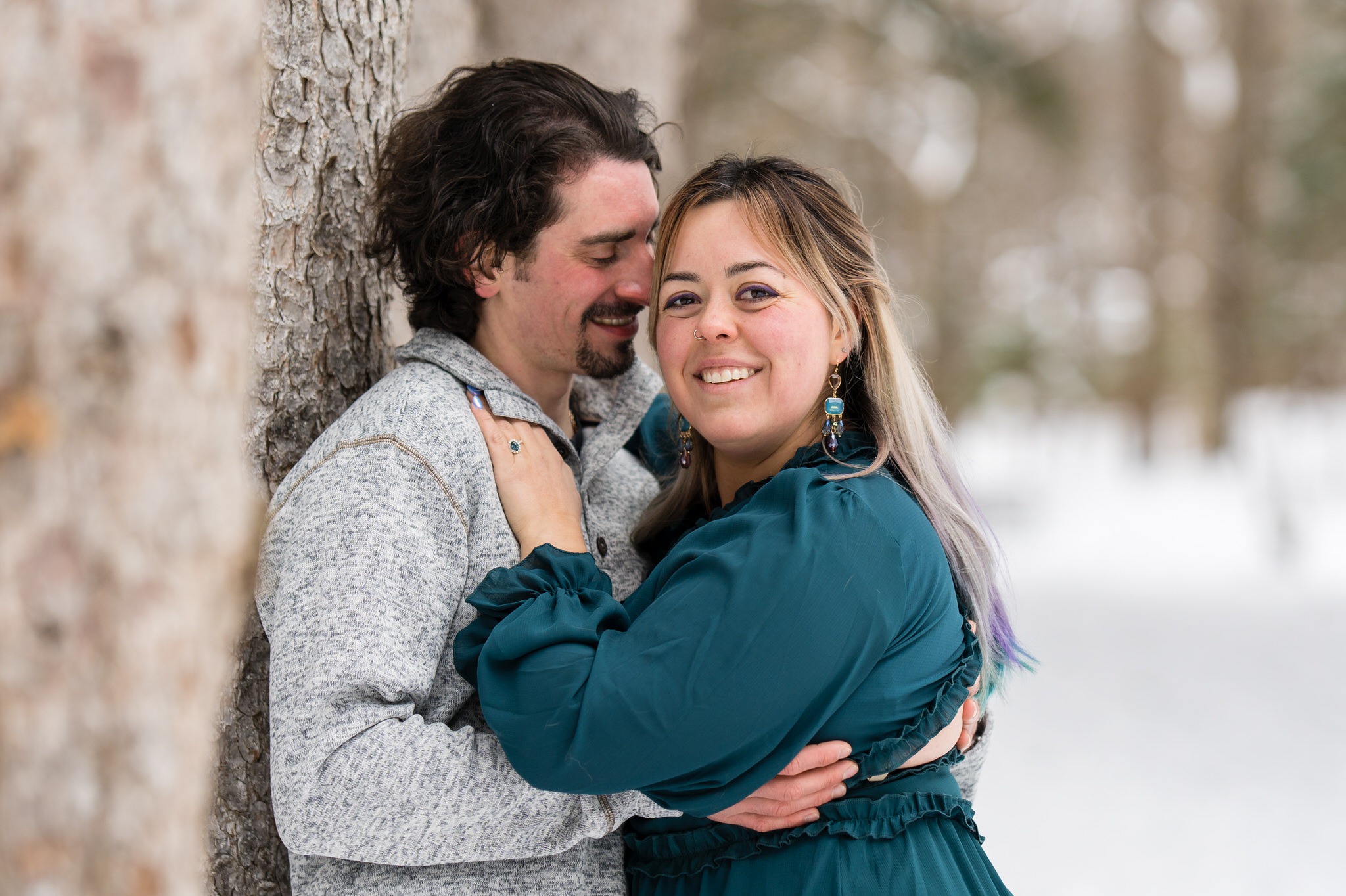 A couple pose leaning on a tree in the snow for their winter engagement session at Stony Creek. 