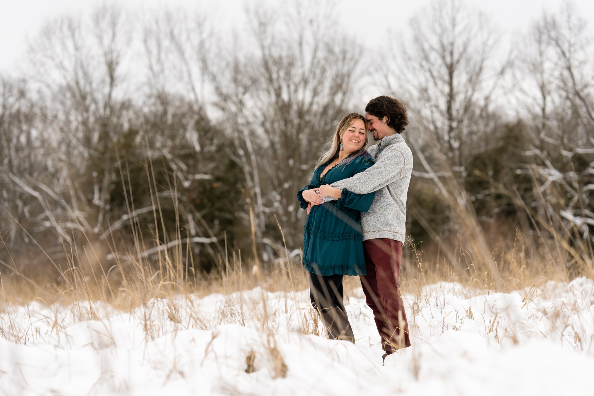 A couple pose in an open field of tall grass and snow for their winter engagement session at Stony Creek. 
