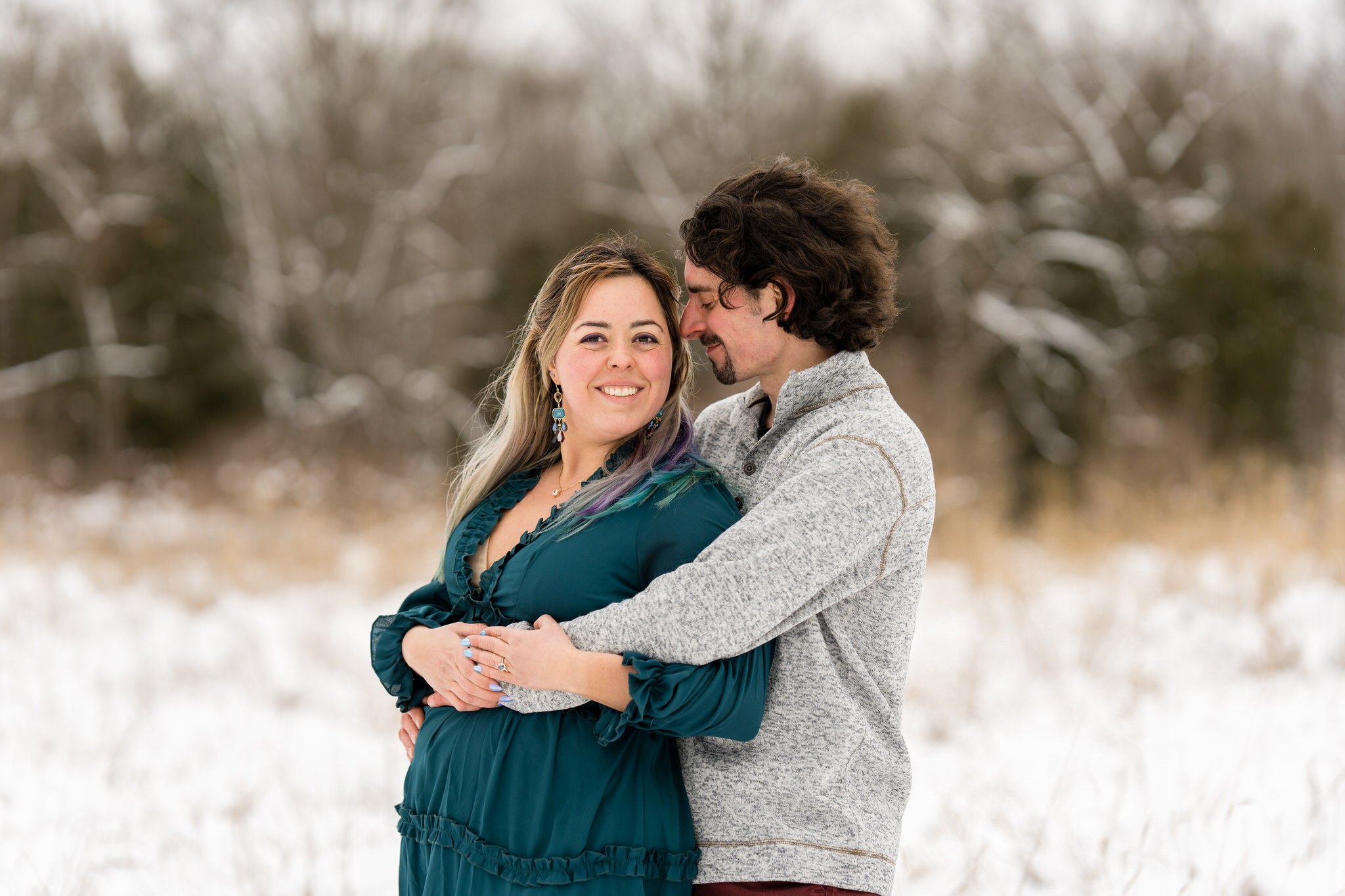 A couple pose in an open field of tall grass and snow for their winter engagement session at Stony Creek. 