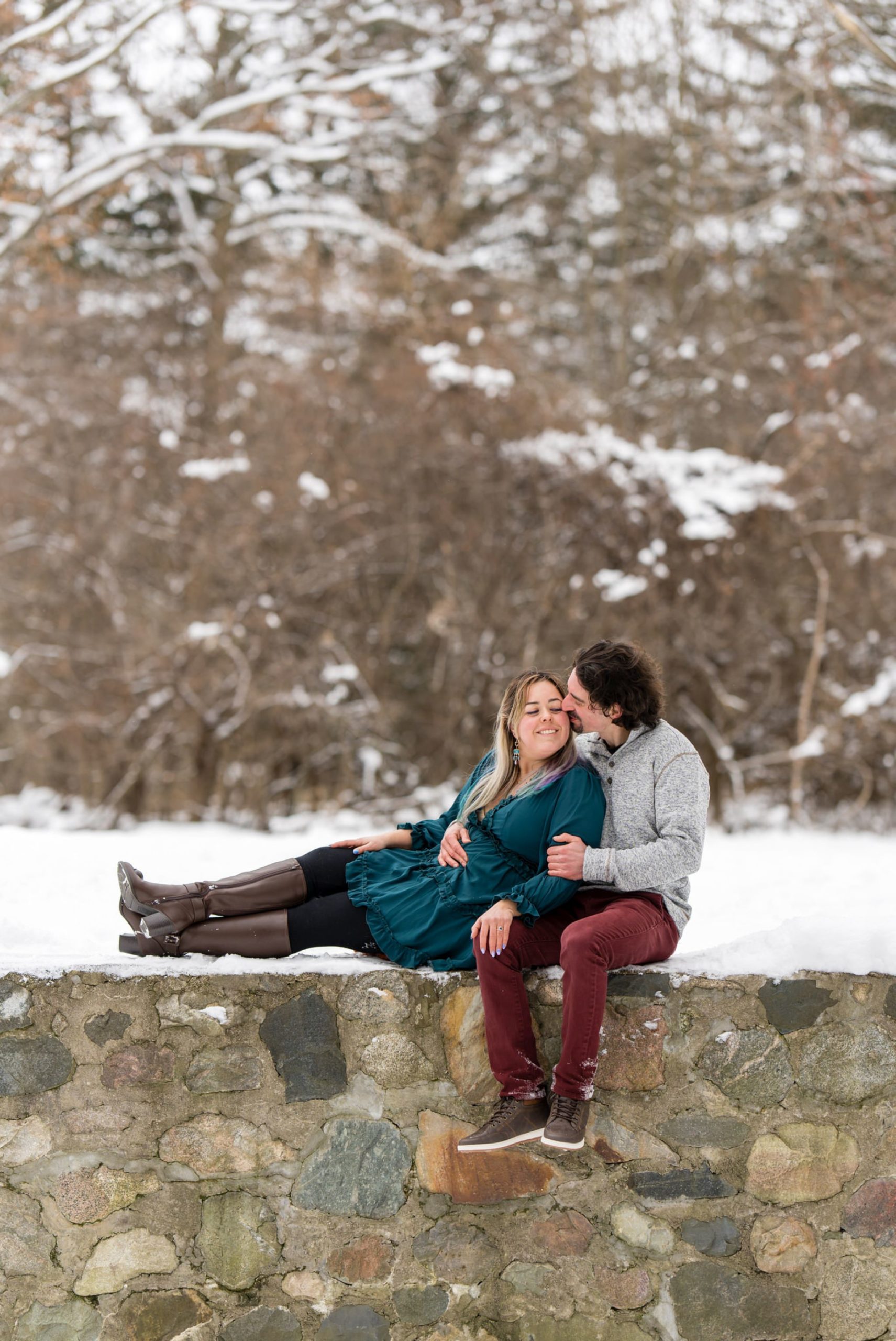 A couple sit on a stone wall at their engagement session at Stony Creek. 