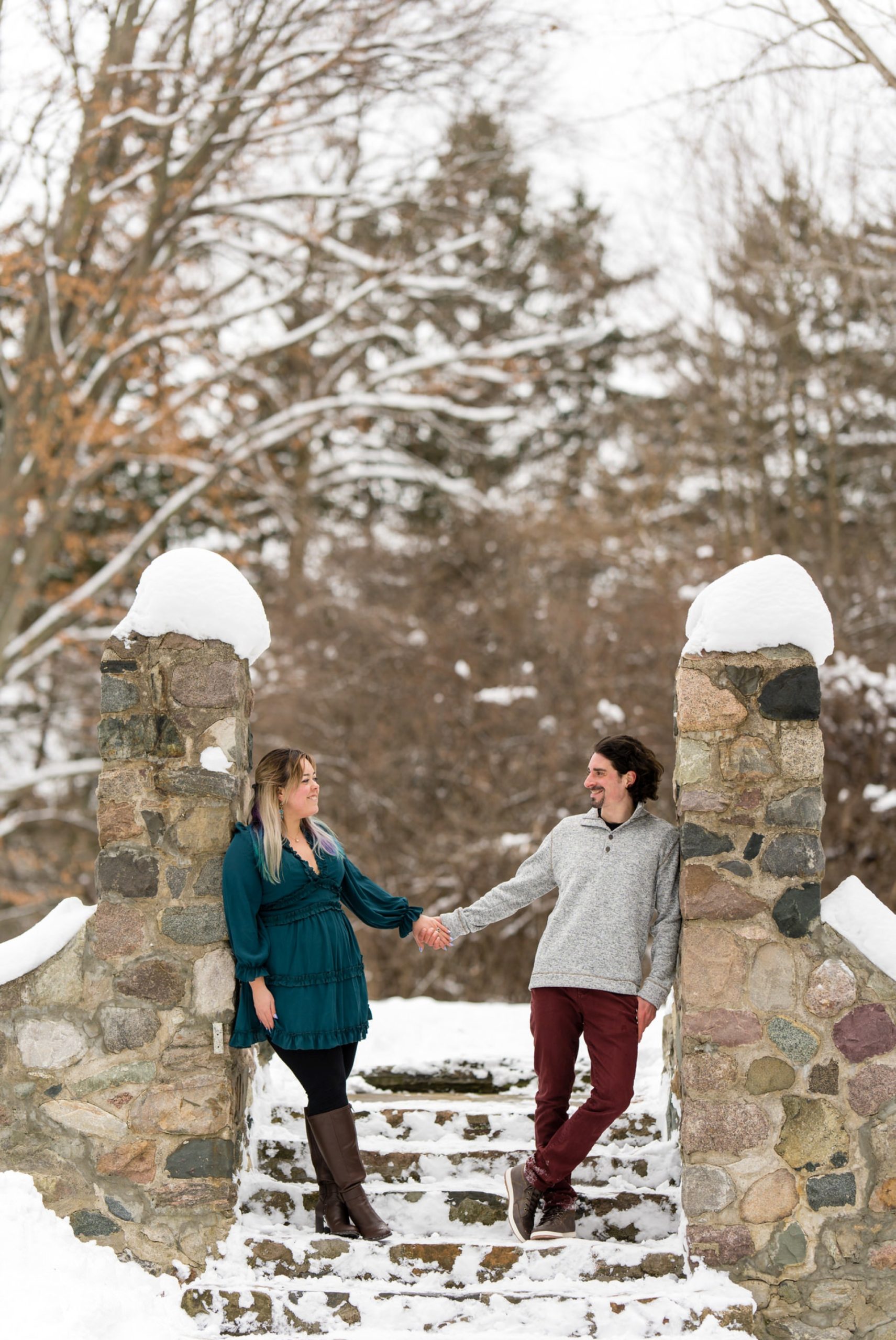 A couple lean on stone wall and hold hands at their engagement session at Stony Creek. 
