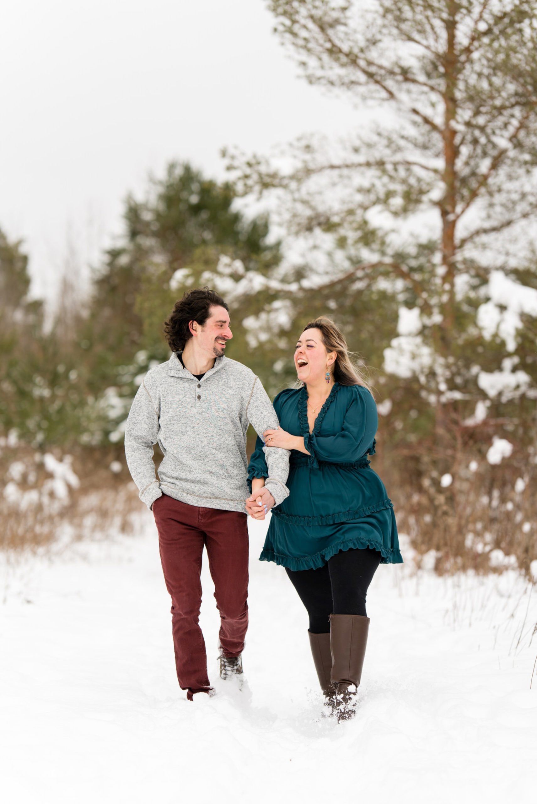 A couple laughs as they walk through the snow at their winter engagement session. 