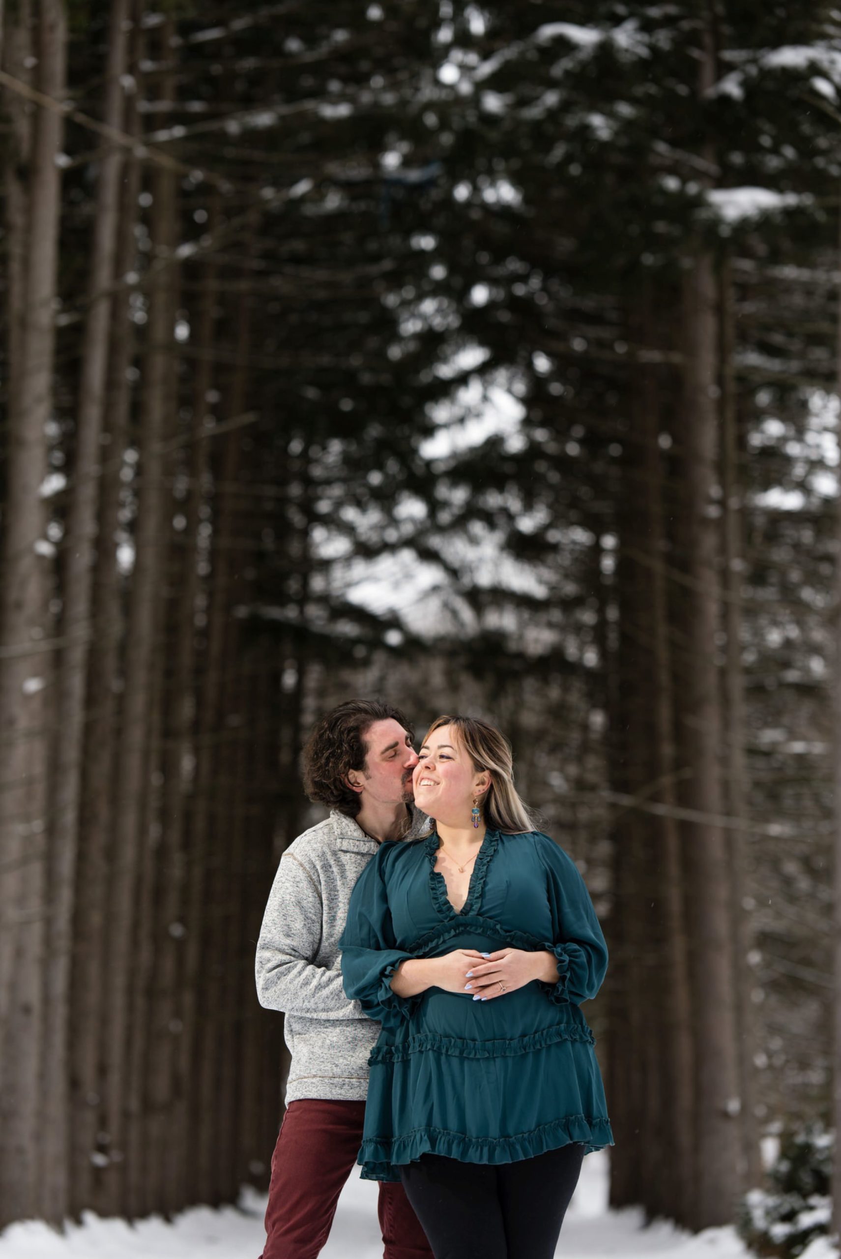 A guy hugs his fiance from behind and kisses her at their winter engagement session at Stony Creek Metropark.