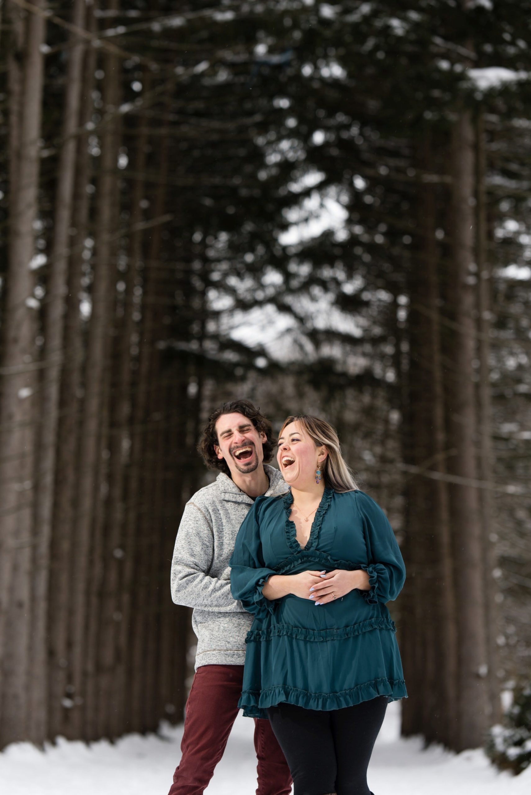 A guy hugs his fiance from behind while they both laugh at their winter engagement session at Stony Creek Metropark.
