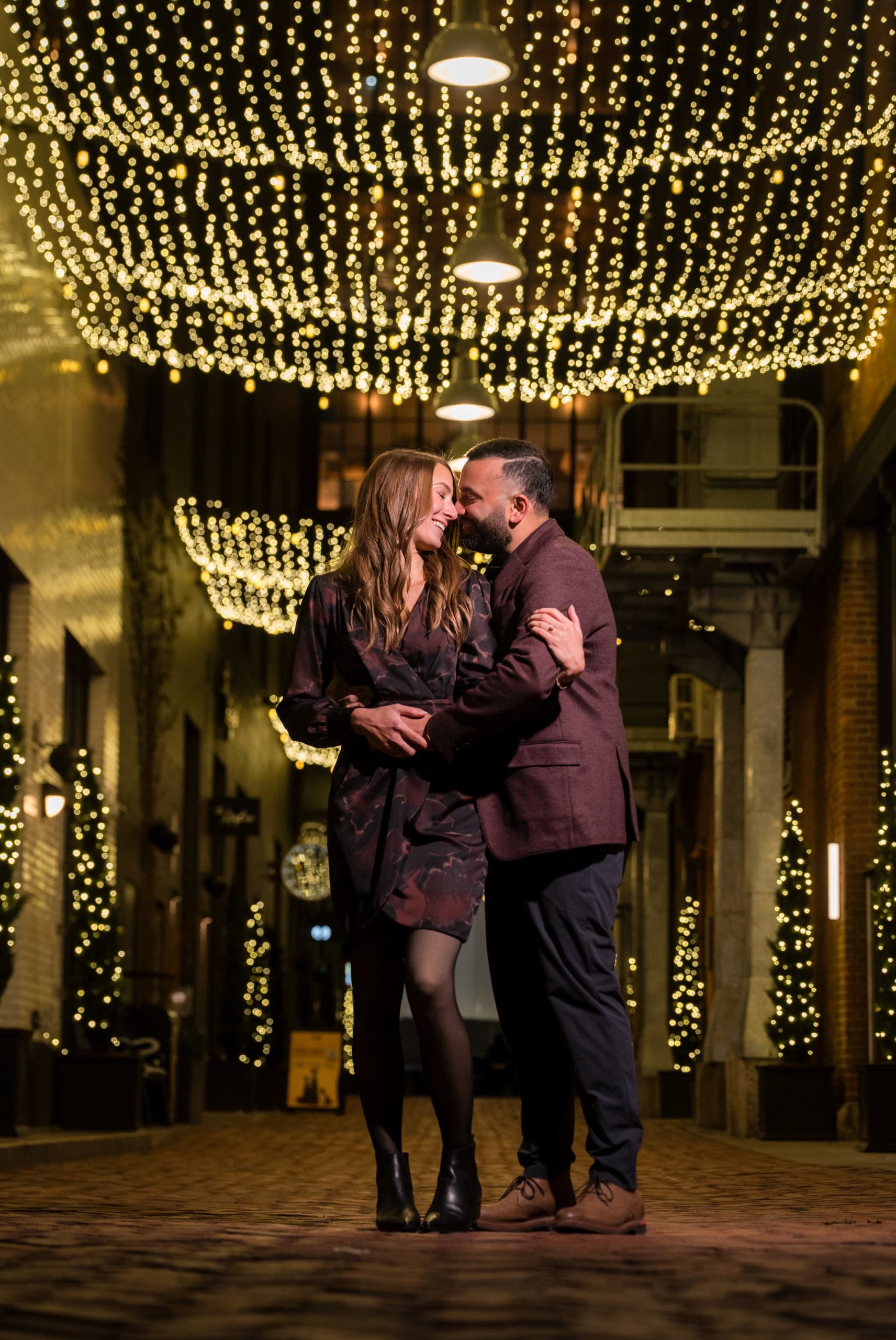 A couple smiles and embraces under the twinkle lights at night  following their Parker's Alley proposal in Detroit. 
