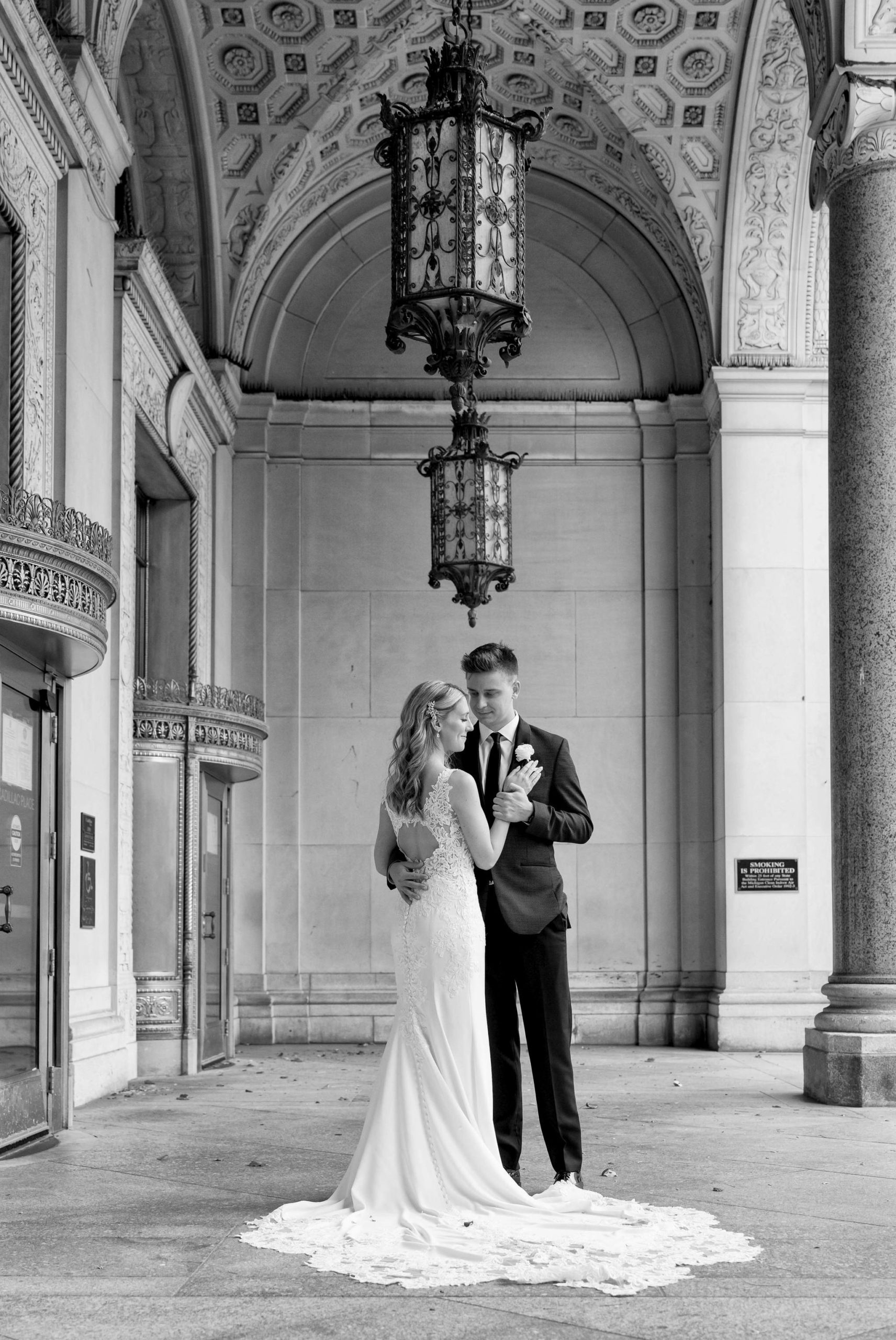 A bride and groom pose elegantly under the arches of the Cadillac Place Apartments on the wedding day.  