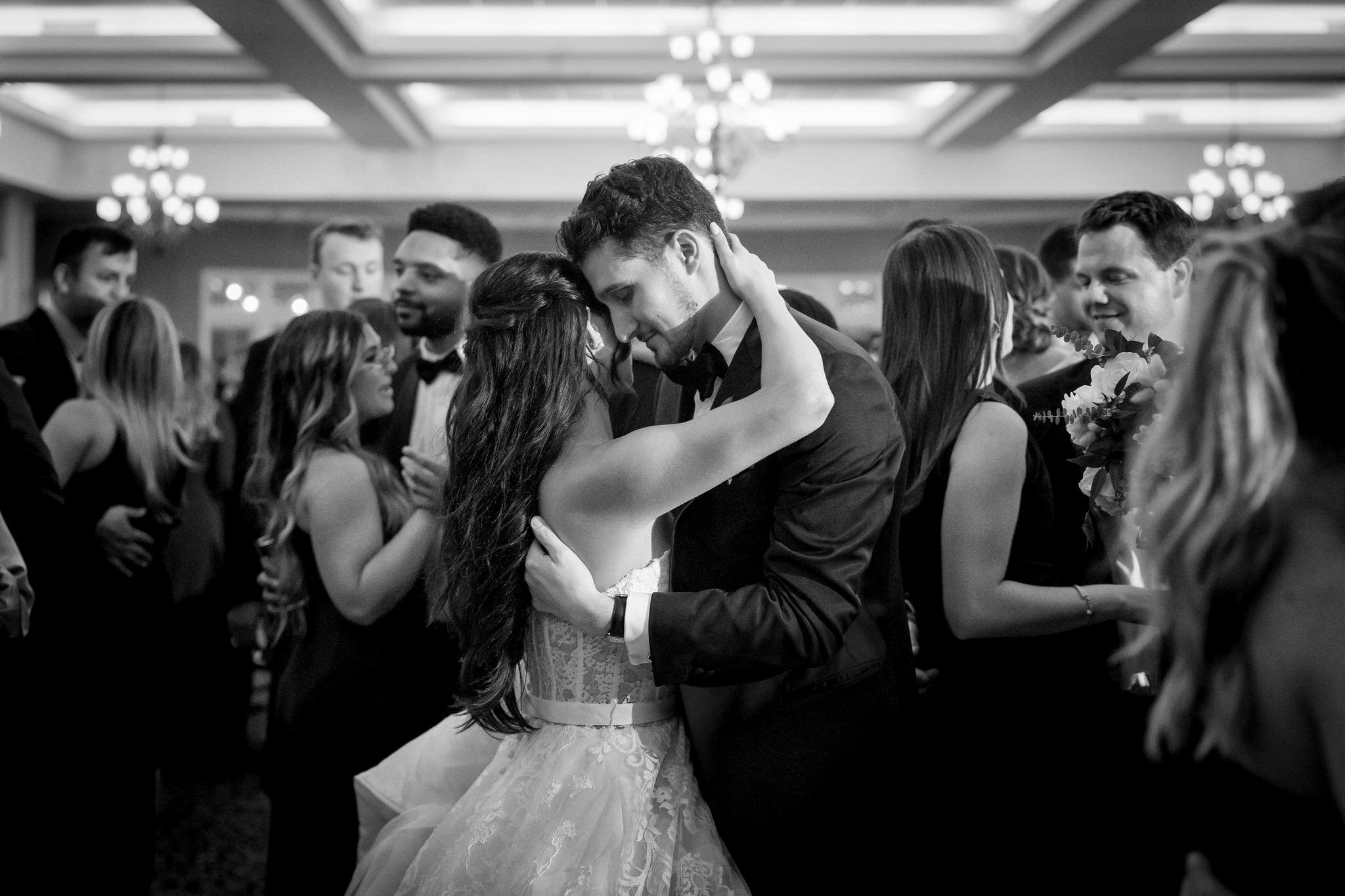 A bride and groom closely embrace while guests dance around them on their wedding day. 