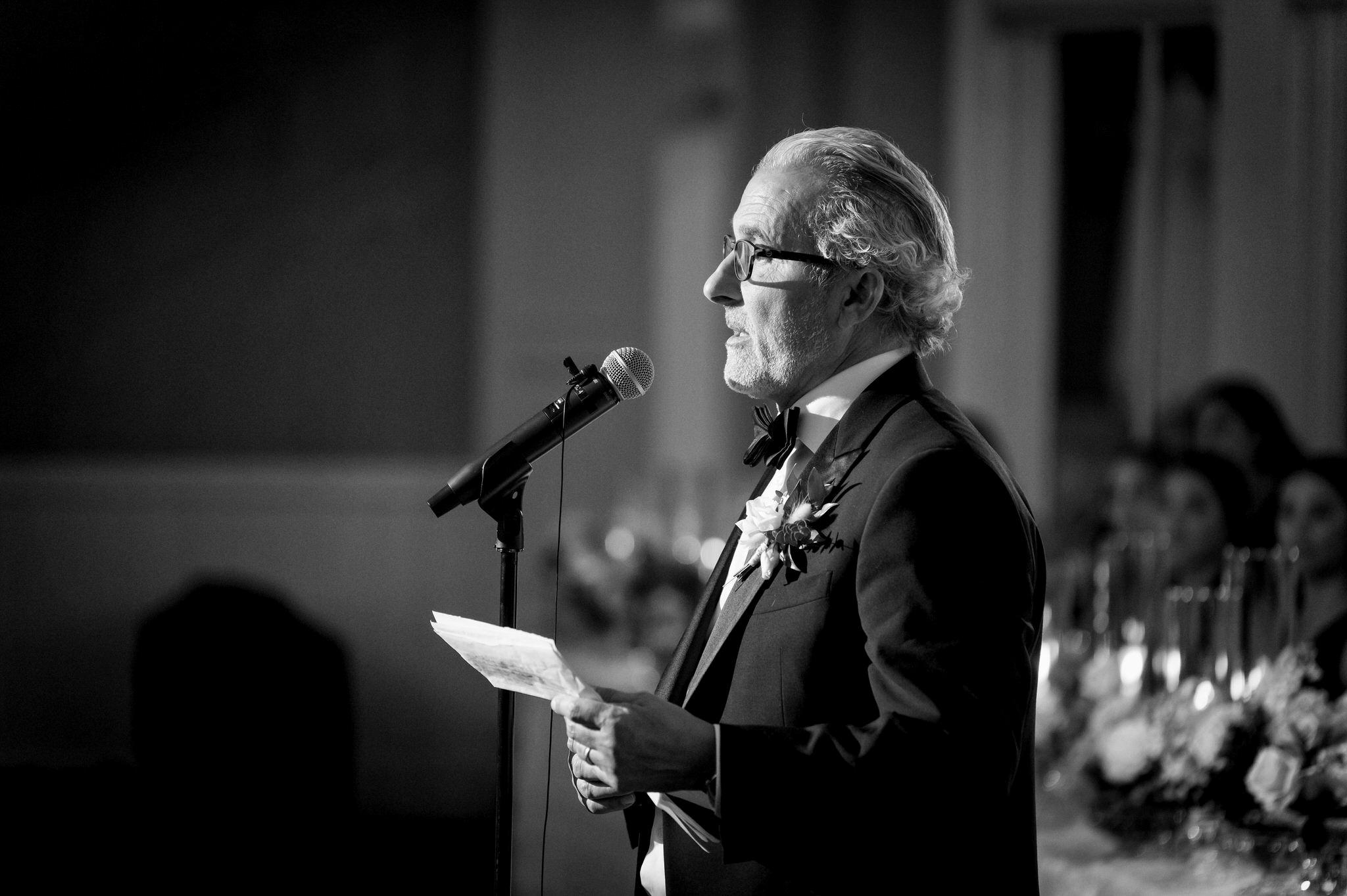 The father of the groom gives a speech while wearing a tuxedo. 