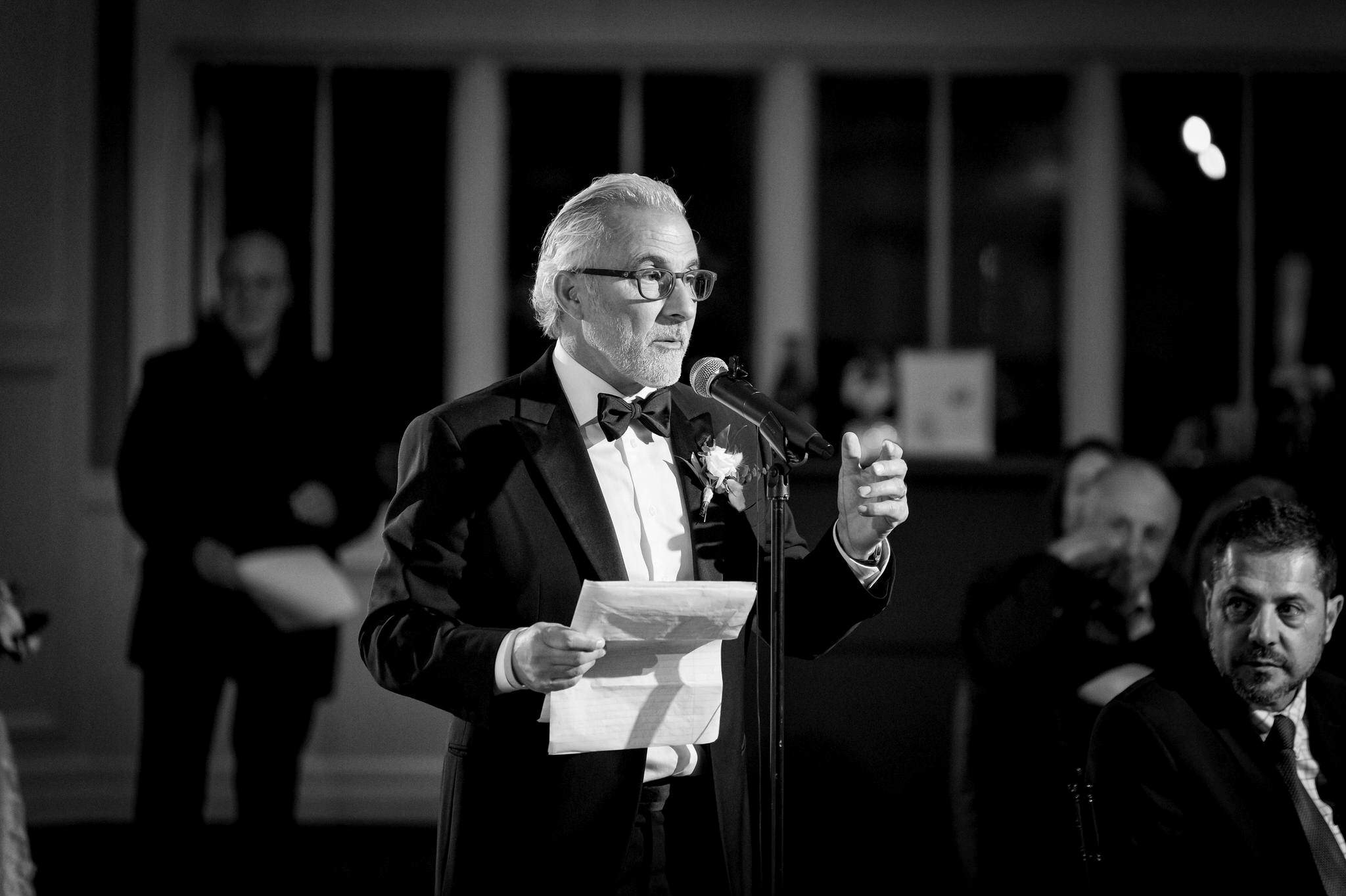 The father of the groom gives a speech while wearing a tuxedo. 