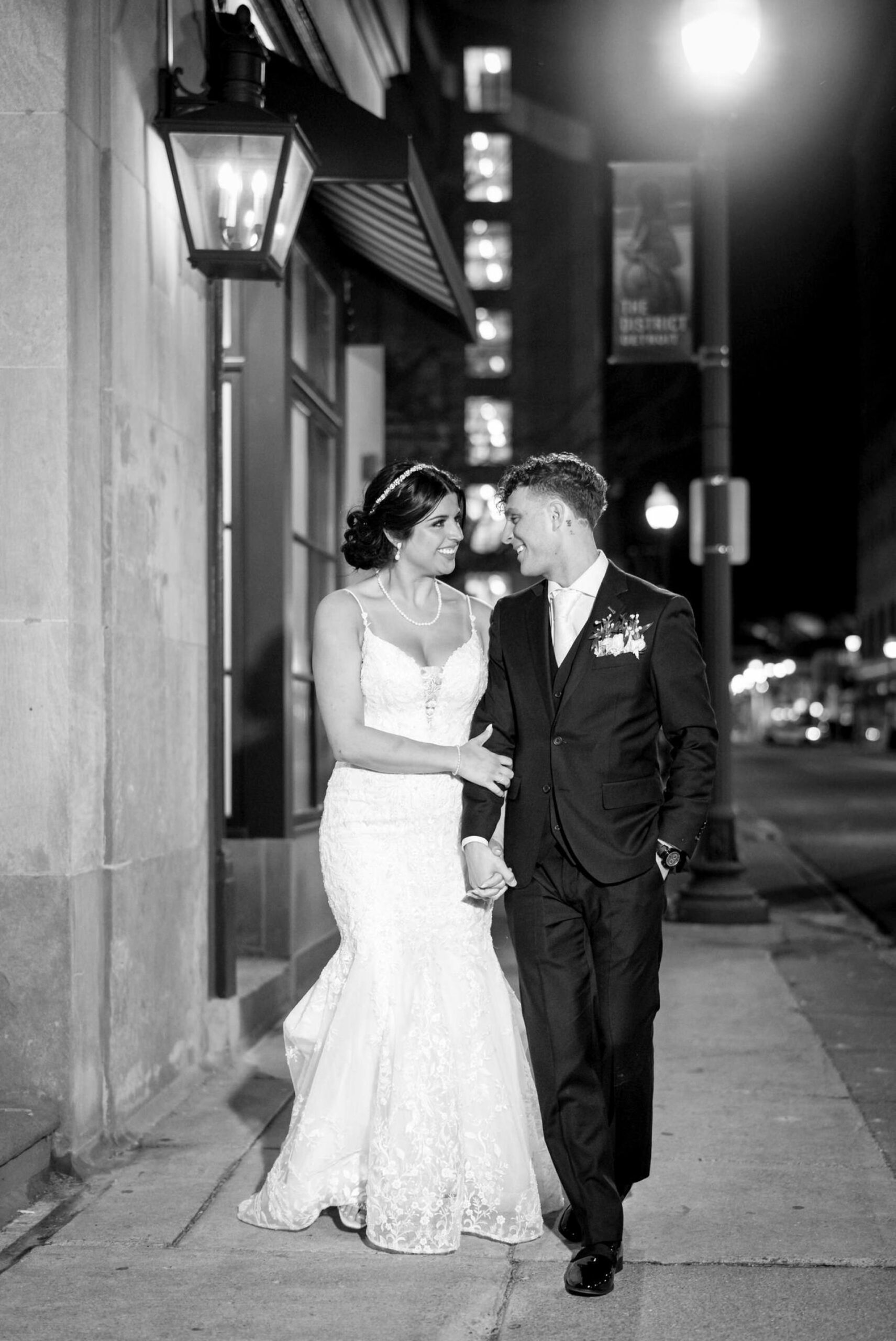 A black and white image of a bride and groom walking outside of their Colony Club Detroit wedding at night. 