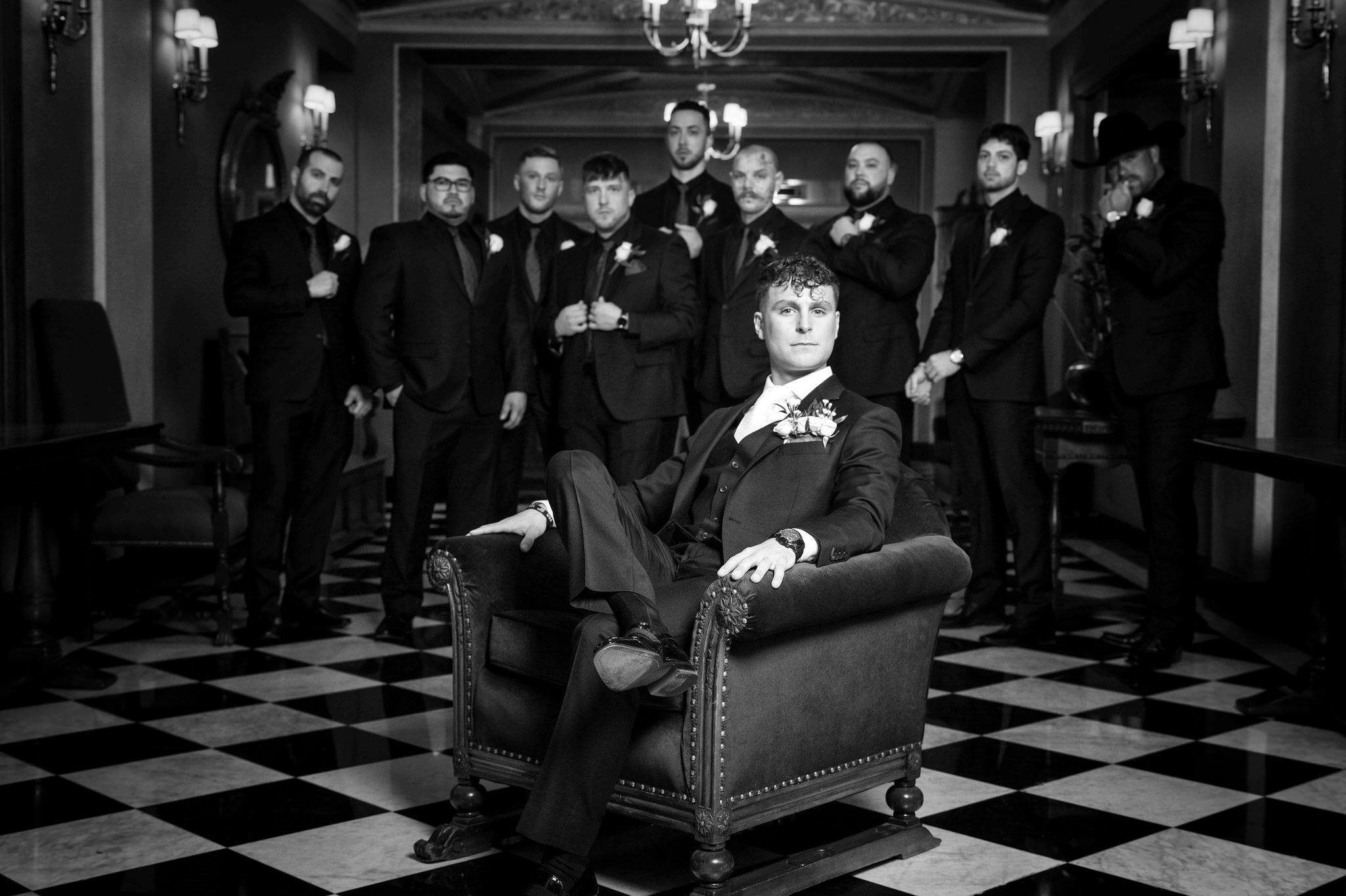A groom sits in a chair and poses with his groomsmen behind him for a portrait on his Colony Club Detroit wedding day. 