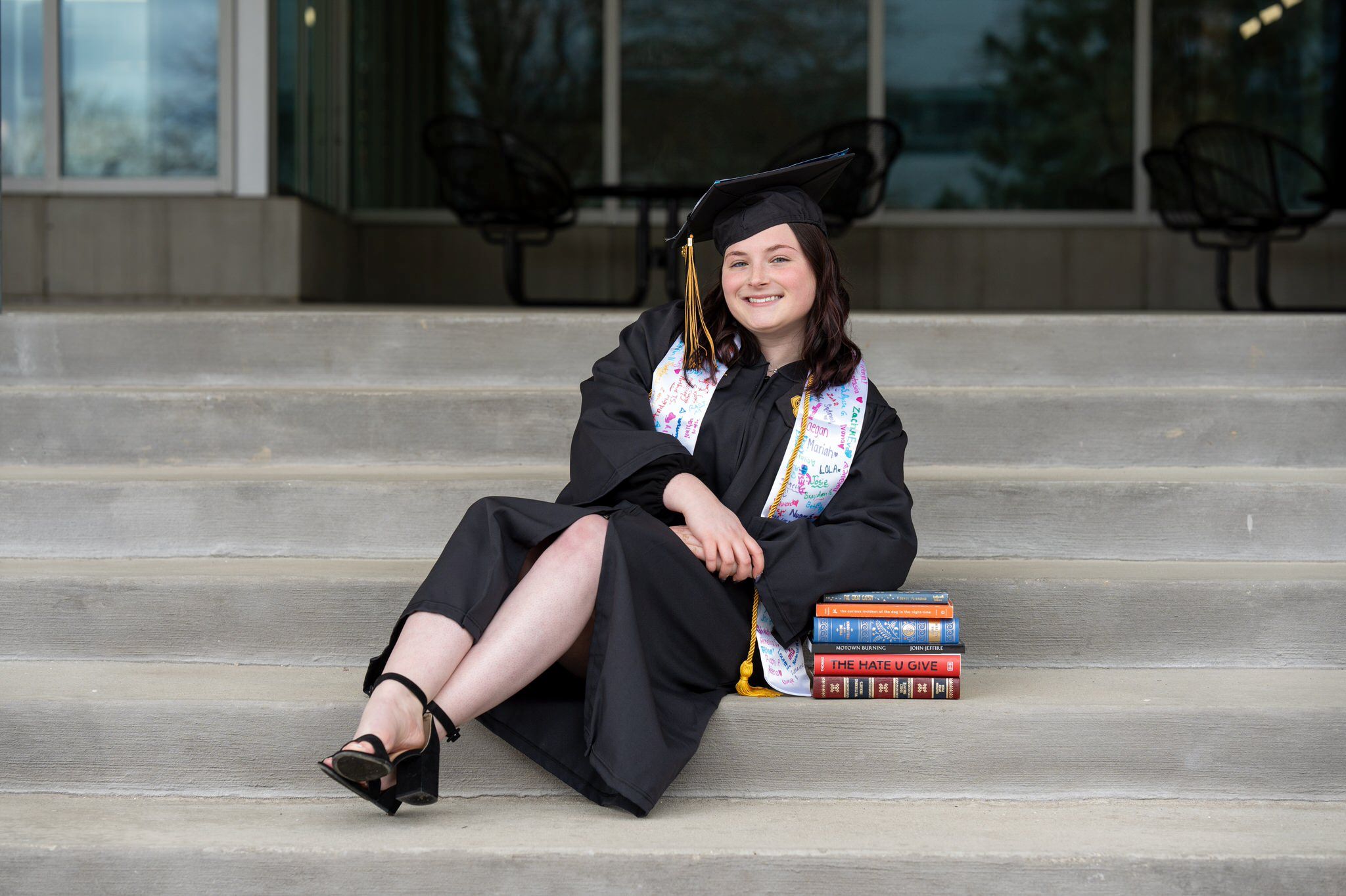 A student leans on a pile of books in graduation regalia
