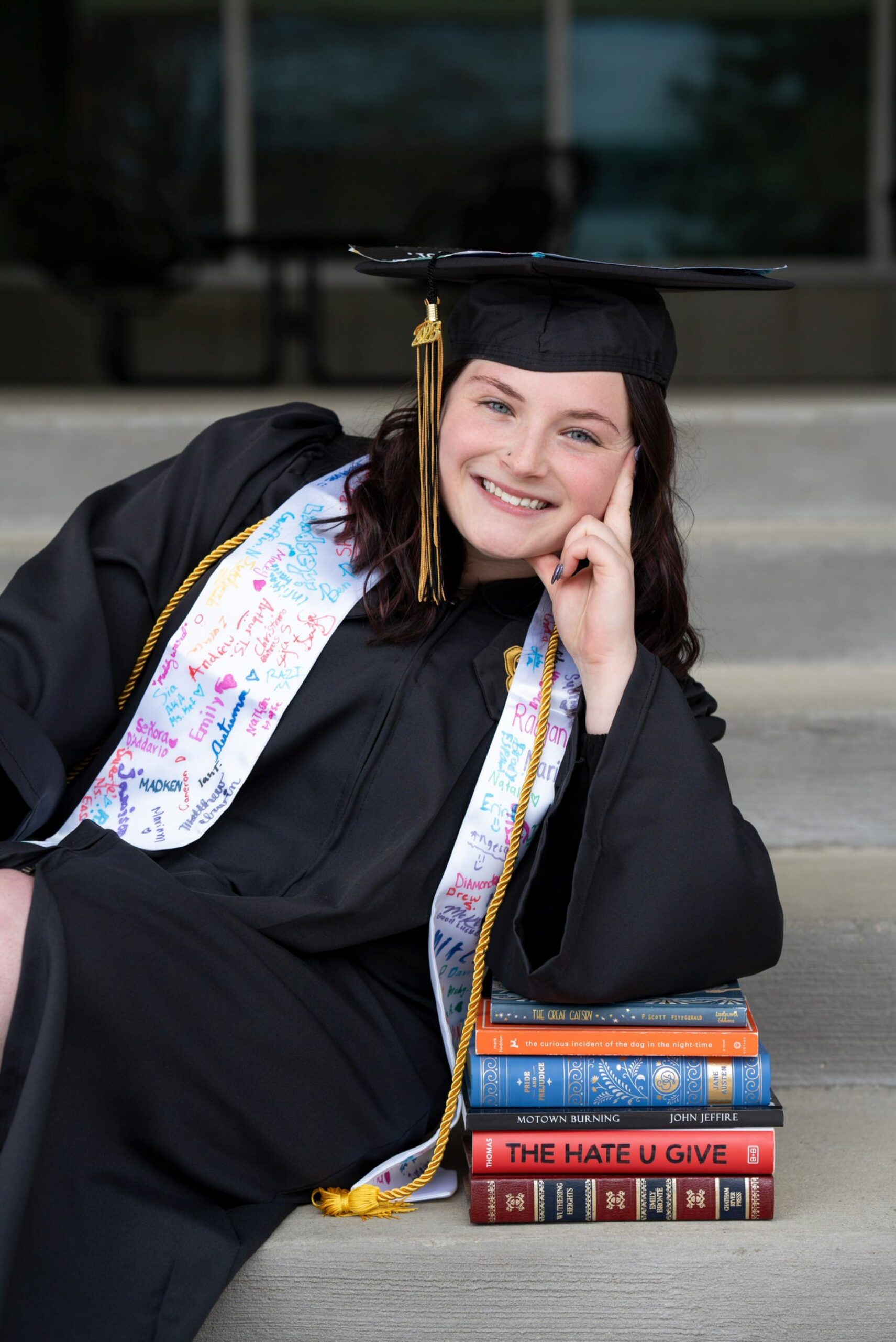 A student leans on a pile of books in graduation regalia