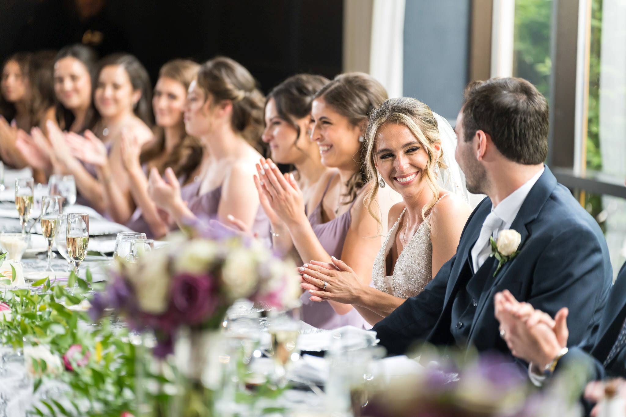 With a tear trickling down her cheek, a bride smiles at her husband during speeches at their Andiamo Showcase wedding in Warren, MI.  