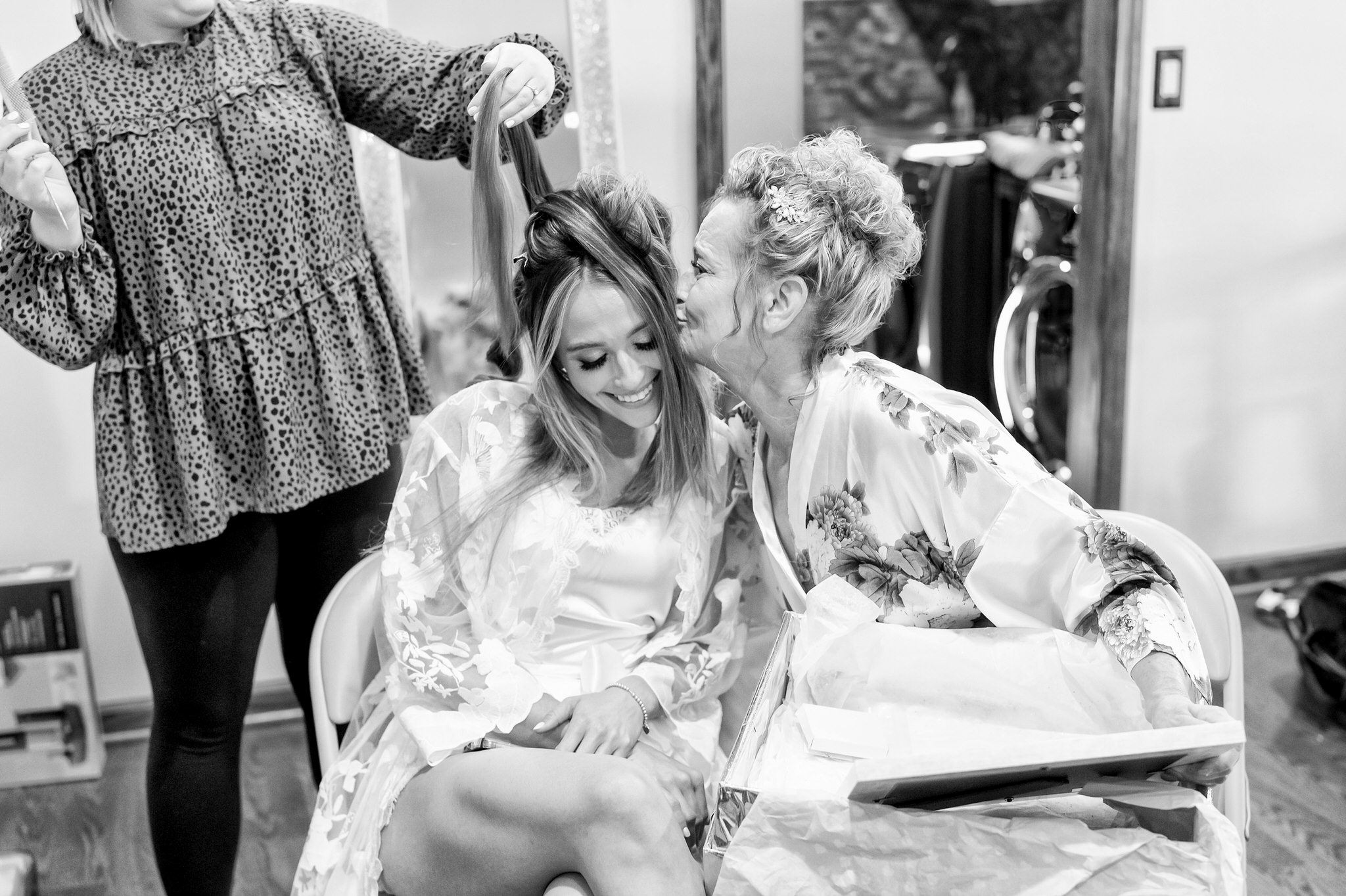 Mom kisses bride, who is getting her hair done on the morning of her wedding.  