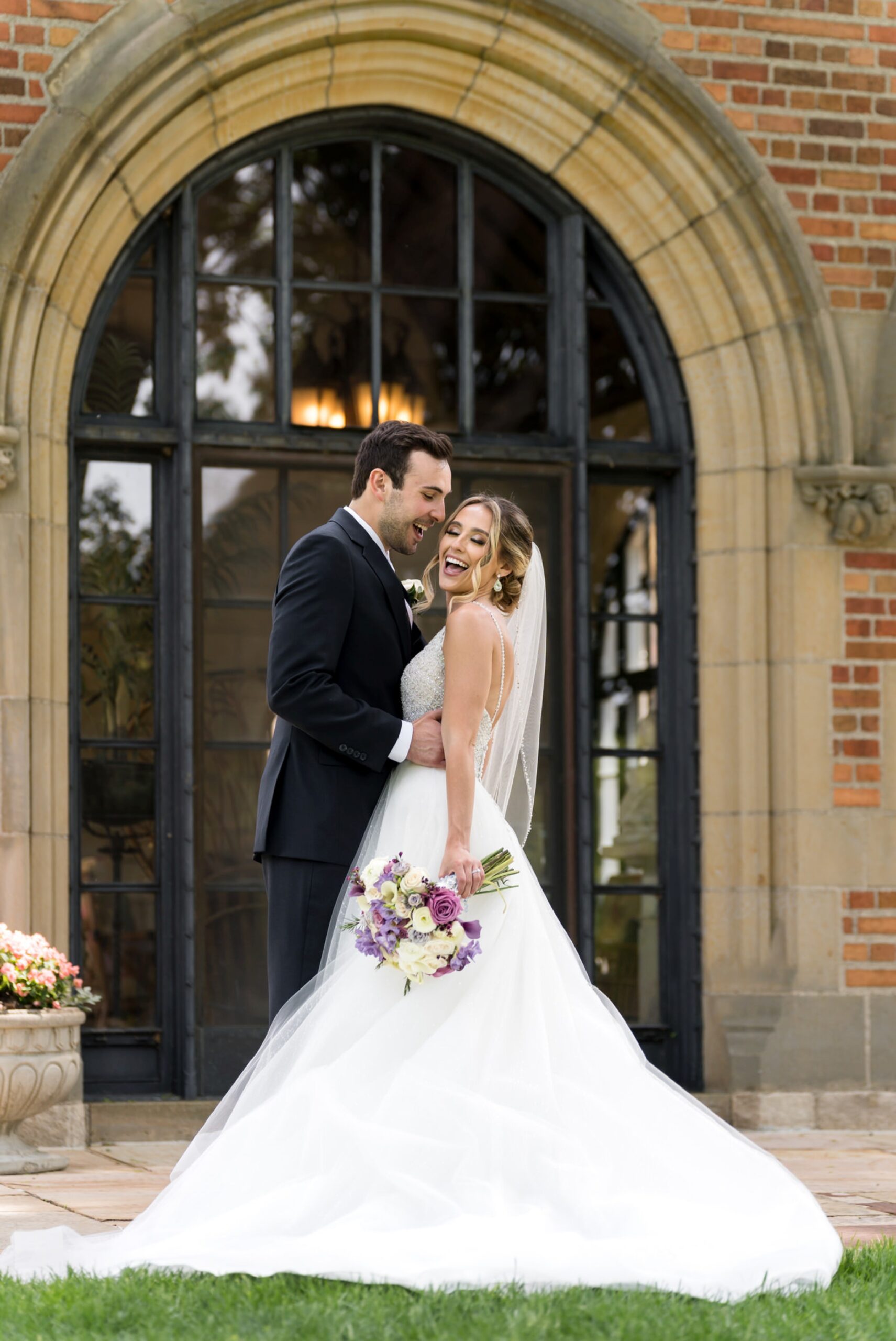 A couple embrace and laugh in front of an arch of windows at their wedding at Meadowbrook Hall.  