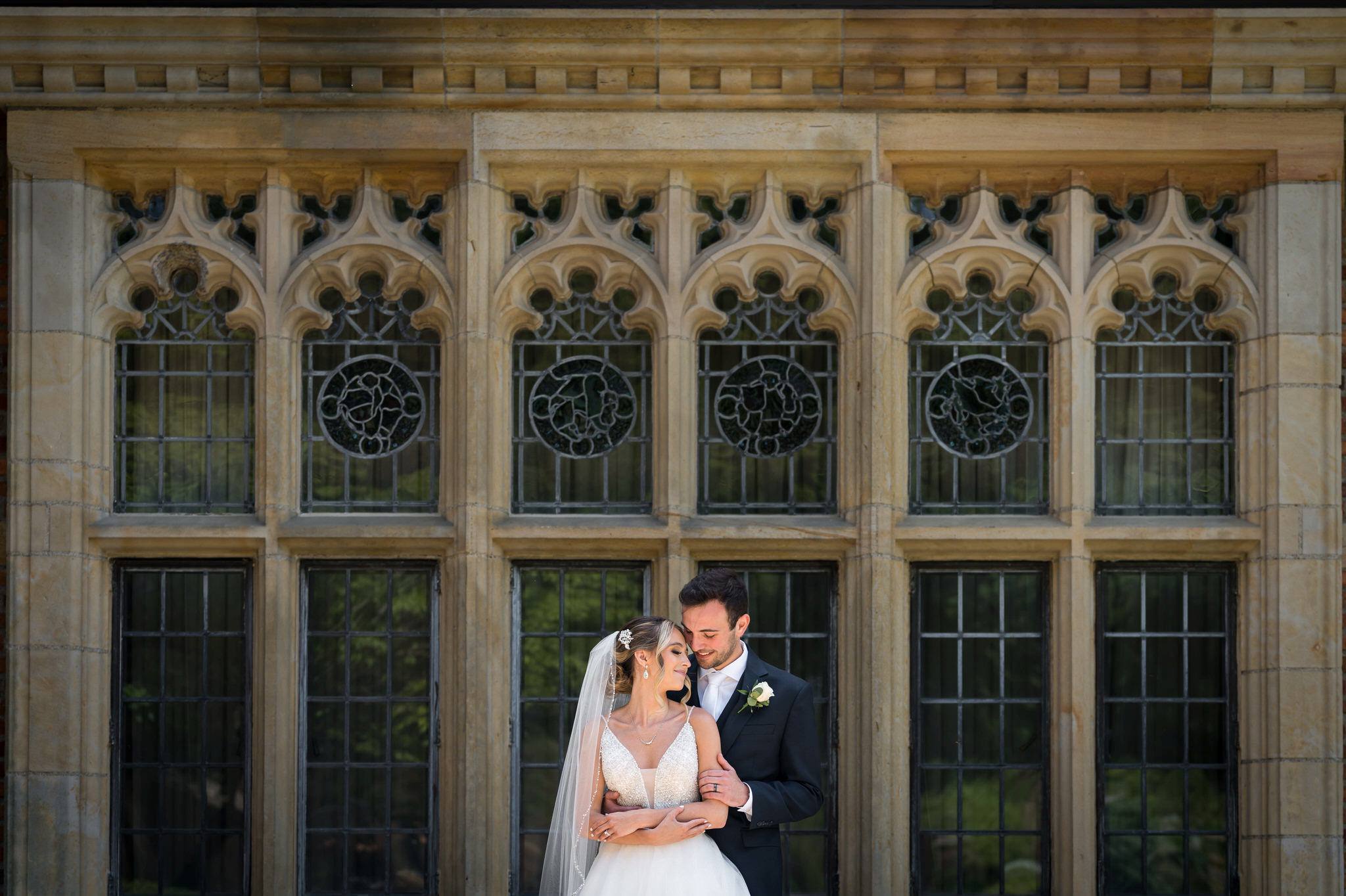 A bride and groom snuggle in front of a wall of windows at their wedding at Meadowbrook Hall.  