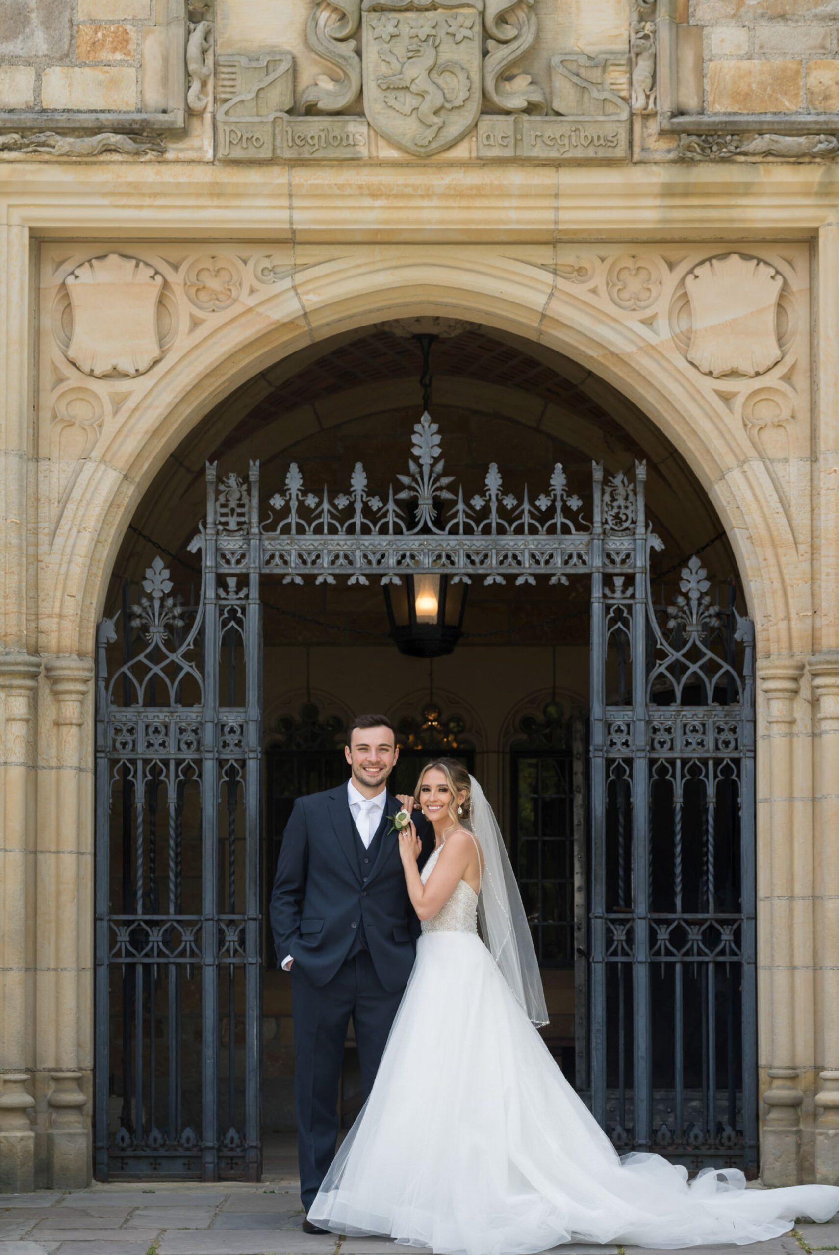 Holding onto his arm, a bride and groom pose in front of the main gate at their wedding at Meadowbrook Hall.  