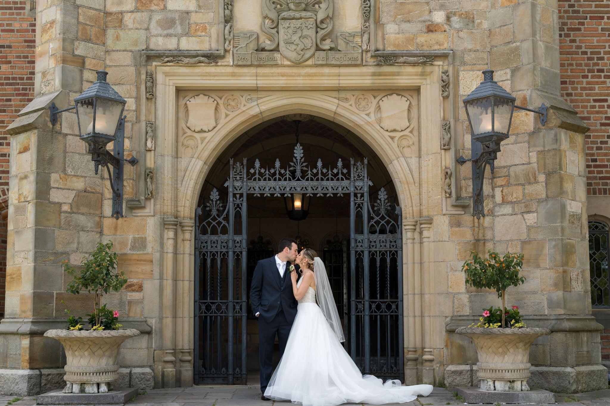 Holding onto his shoulder, a bride and groom pose in front of the main gate at their wedding at Meadowbrook Hall.  