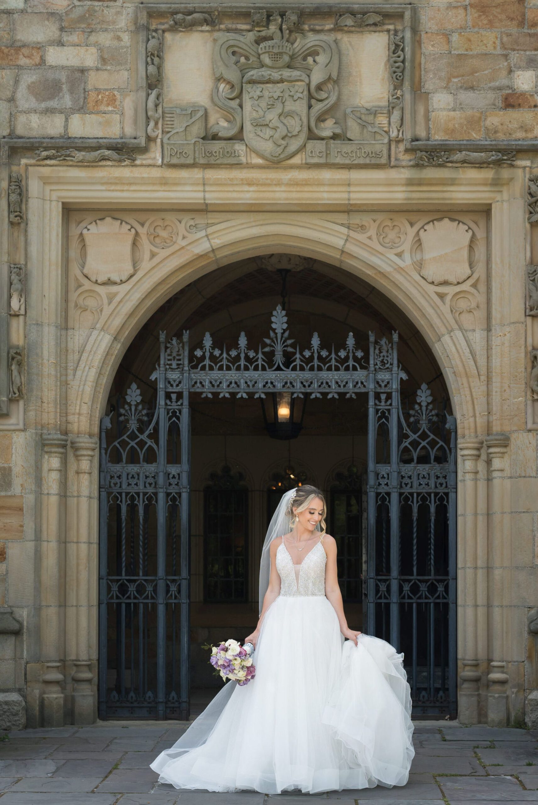 Holding a purpose and white bouquet in one hand and her dress in another, a bride poses in front of the main gate at her wedding at Meadowbrook Hall. 